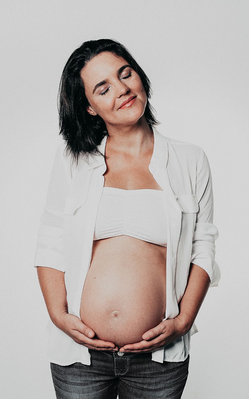 Schwangere Frau steht vor weissem Hintergrund im Fotostudio. Sie hält ihren Bauch mit geschlossenen Augen und lächelt.