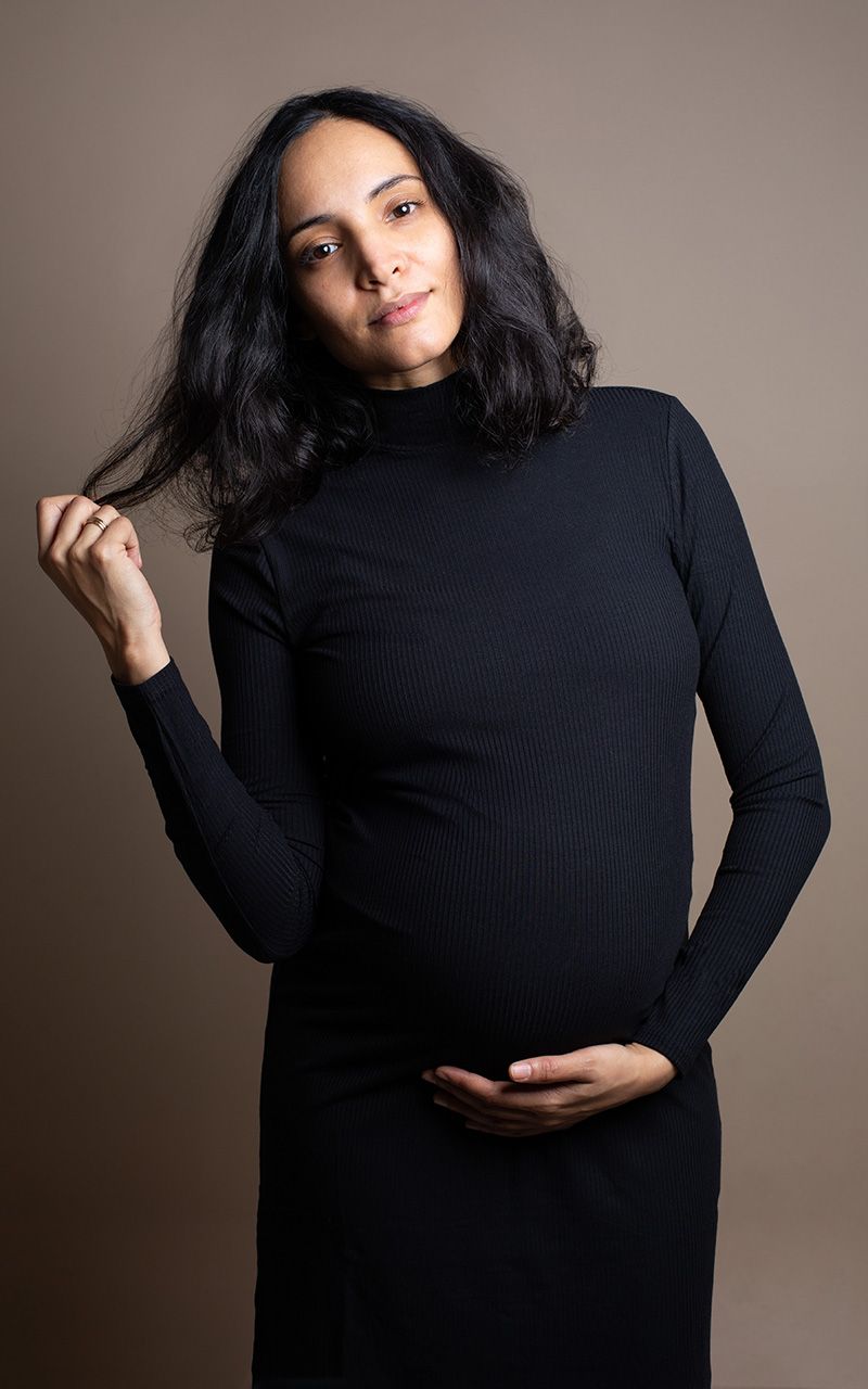 Schwangere Frau trägt ein schwarzes Kleid, sie fasst sich in ihr schwarzes Haar.
