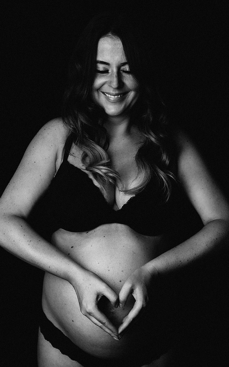 Schwangere Frau in schwarzen Dessous vor schwarzem Hintergrund die mit ihrem Händen ein Herz vor dem Bauch formt.