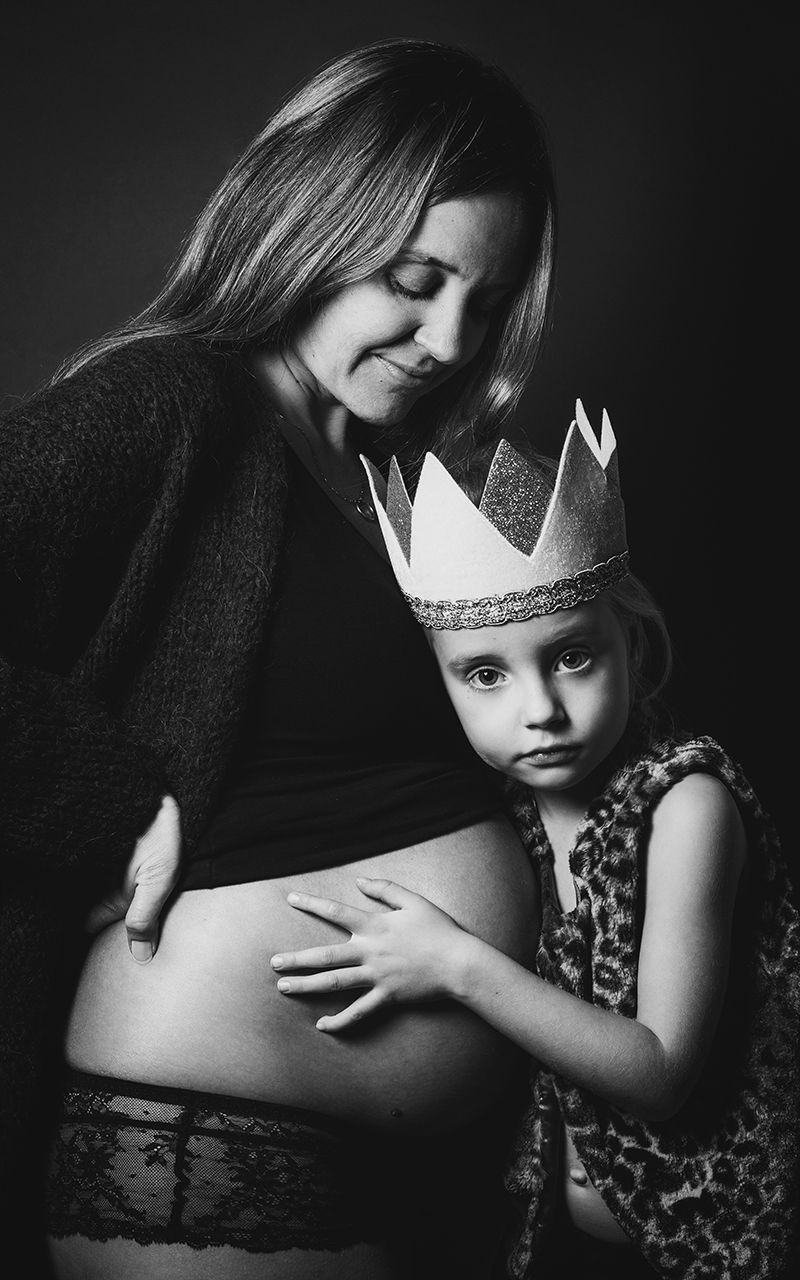 Mutter mit Babybauch steht mit ihrer Tochter im Fotostudio. Die Tochter trägt eine Krone und eine Veste.
