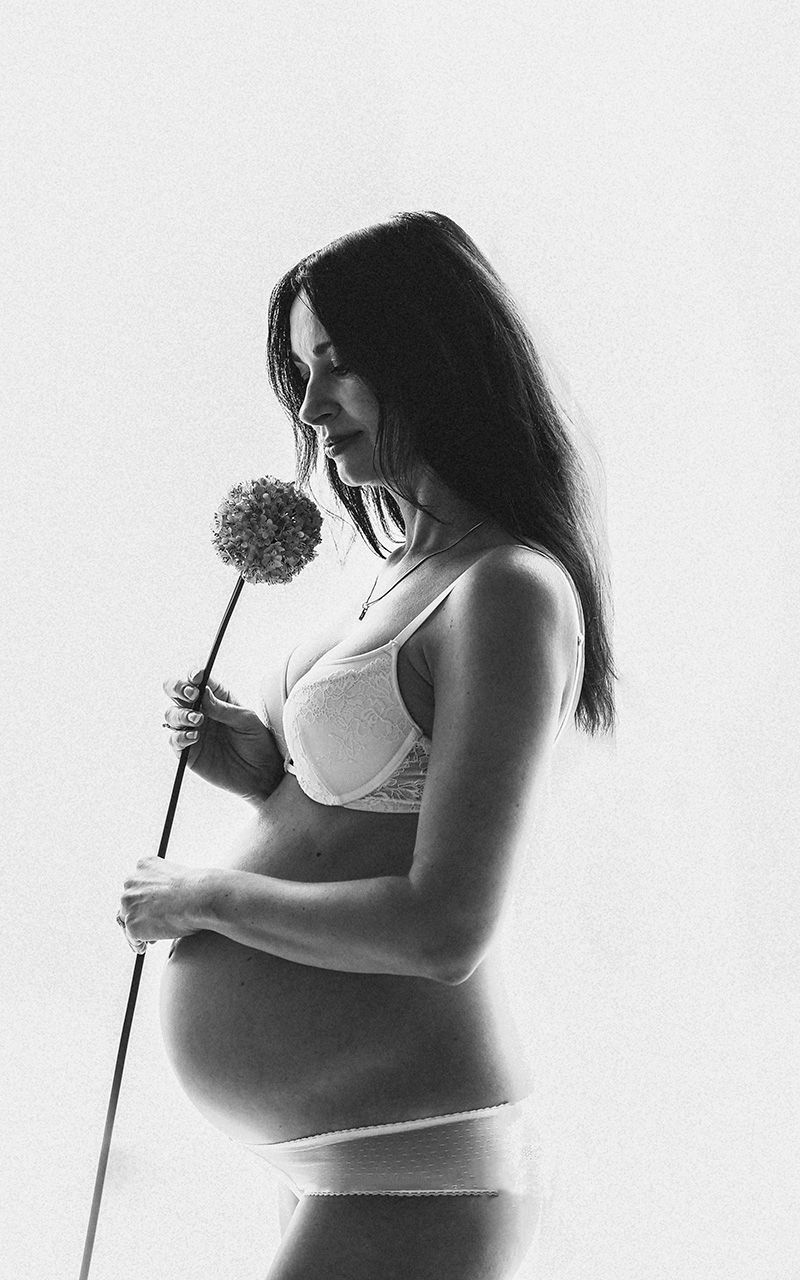 Schwangere Frau steht im Studio in Unterwäsche und riecht an einer Blume.