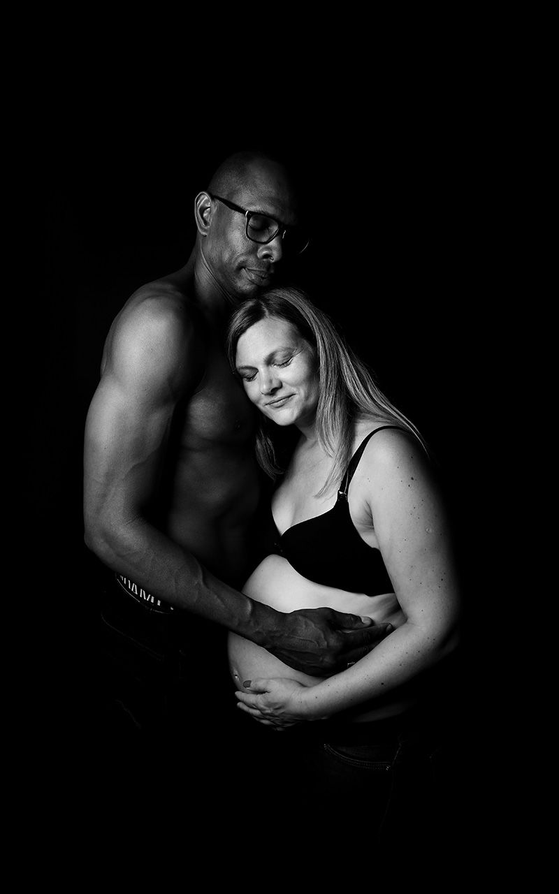 Paar umarmt sich liebevoll im Fotostudio, der Mann hält den Babybauch der Frau
