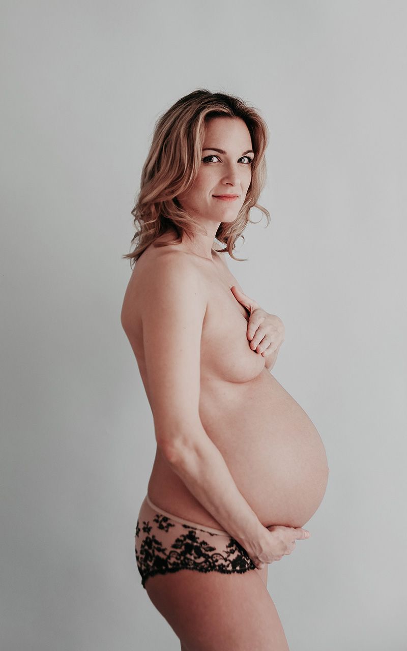 Schwangere Frau steht vor hellblauem Hintergrund im Fotostudio, oben trägt sie nichts und hält ihre Hand auf die Brust.