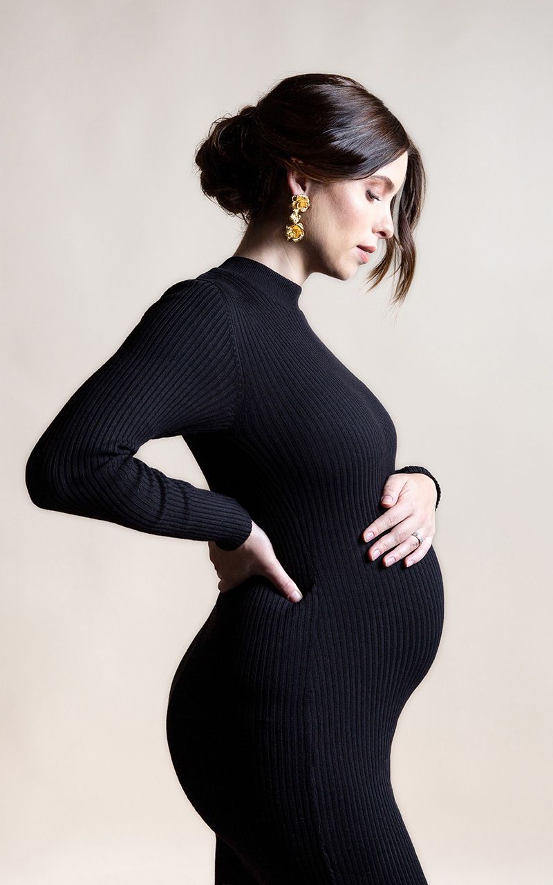 Schwangere Frau steht im Fotostudio und hält mit ihren Händen den Bauch. Sie trägt ein schwarzes Kleid.