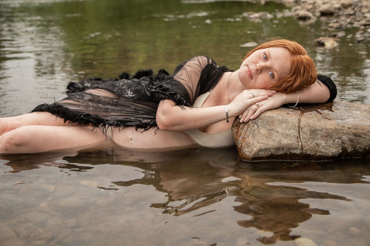 Red-haired woman in a black dress lying peacefully on a rock in shallow river water.