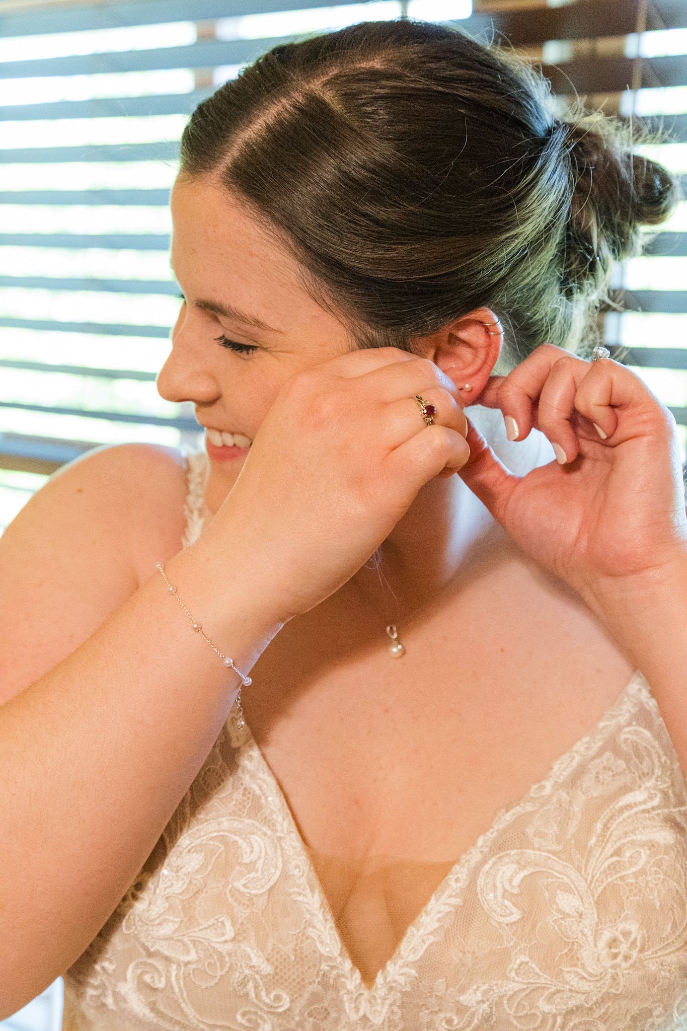 Smiling bride in lace dress putting on pearl earrings near a window before her wedding.