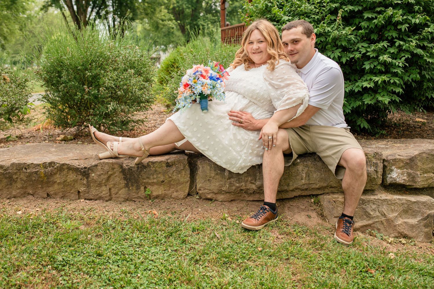 Casual wedding couple sitting on stone wall in garden, bride holding colorful bouquet in short lace dress.