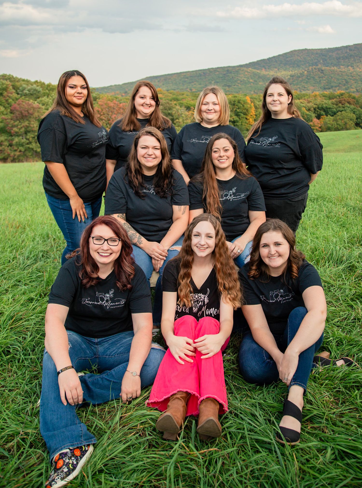 Group of nine women in matching black shirts posing outdoors in a green field with scenic mountain views.