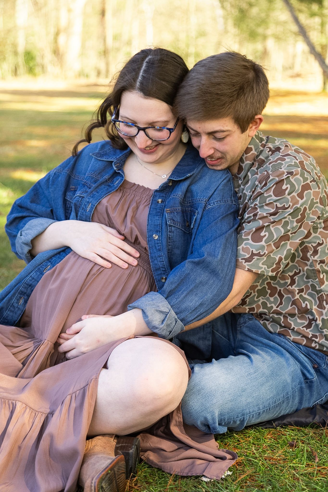 Couple sitting on grass in a sunny park, man embracing pregnant woman from behind, both cradling her baby bump.