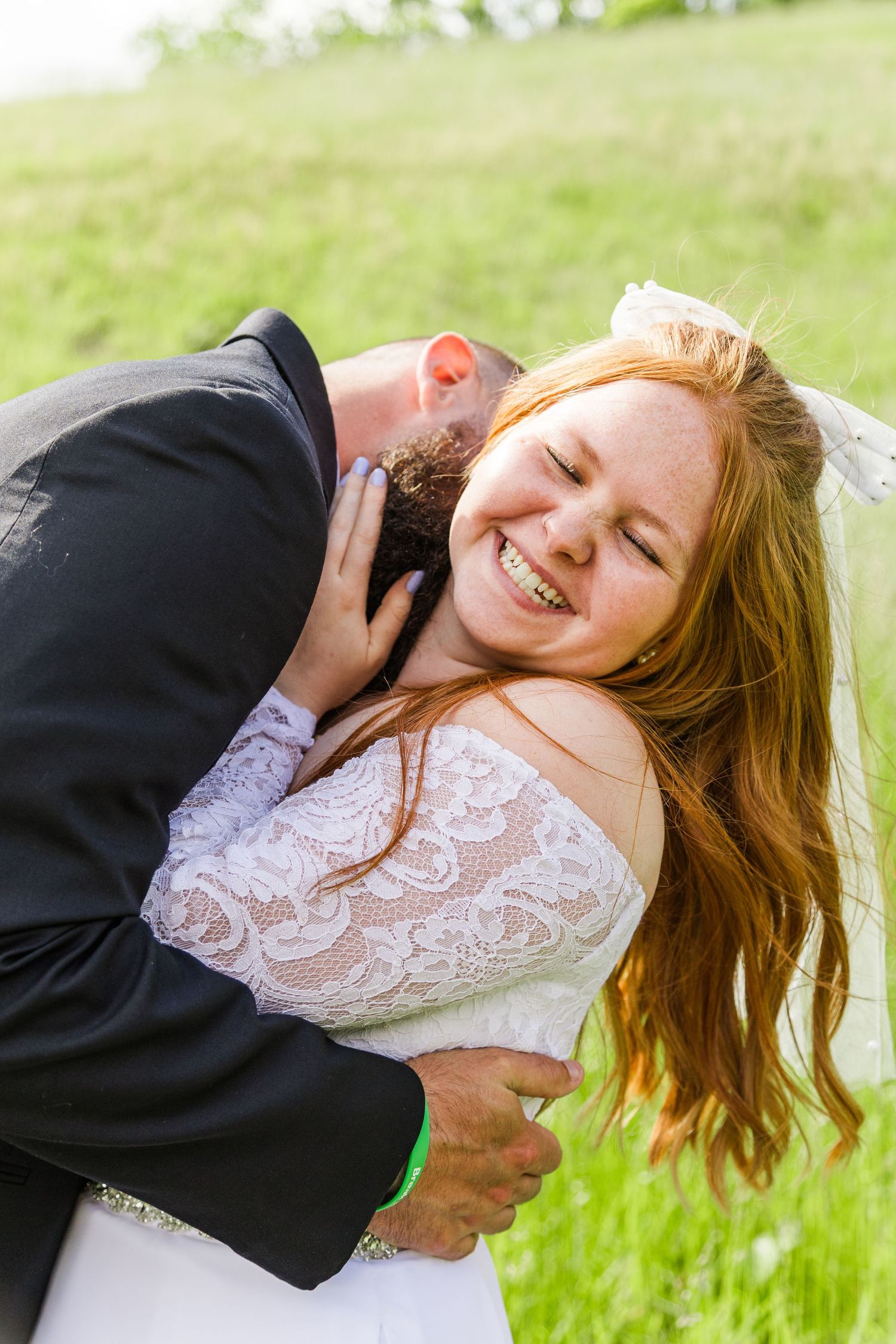Joyful redhead bride laughs as groom kisses her neck in an outdoor wedding portrait.