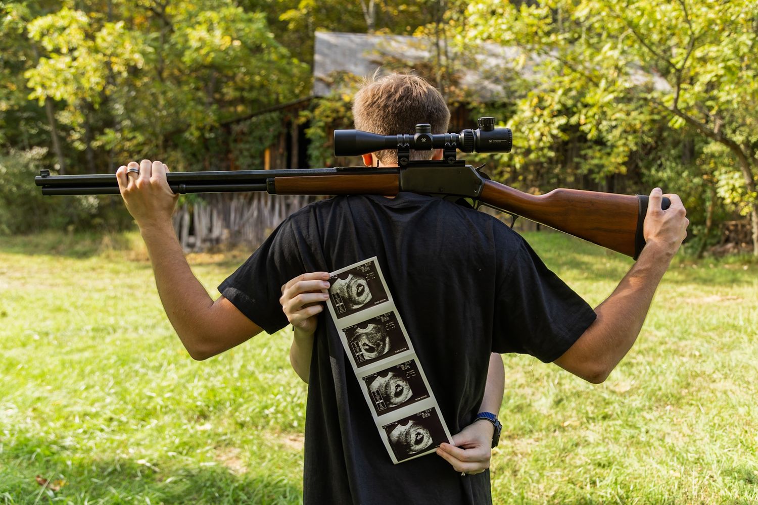 Man holding a rifle across his shoulders while displaying ultrasound photos in a rural outdoor setting.