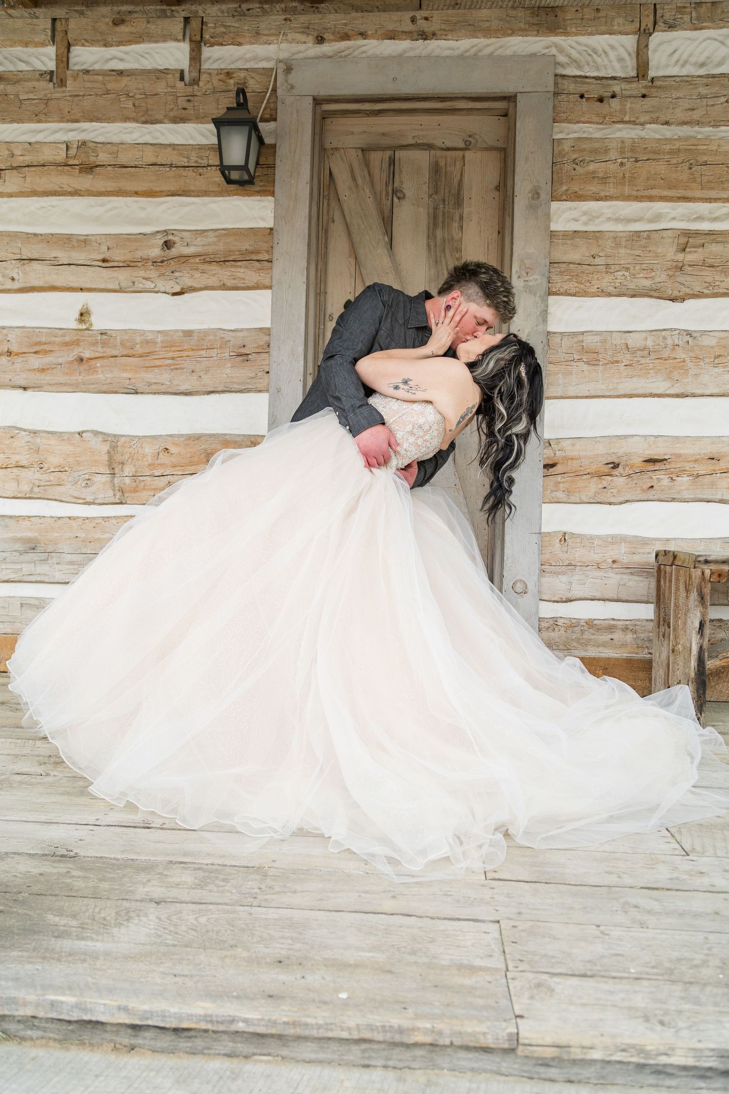 Groom dips bride in full ballgown for romantic kiss on rustic log cabin porch.
