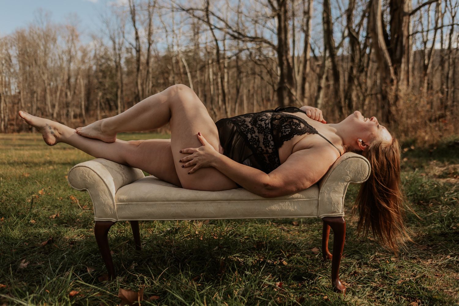 Woman in black lace bodysuit reclines on vintage chaise lounge in outdoor autumn setting.