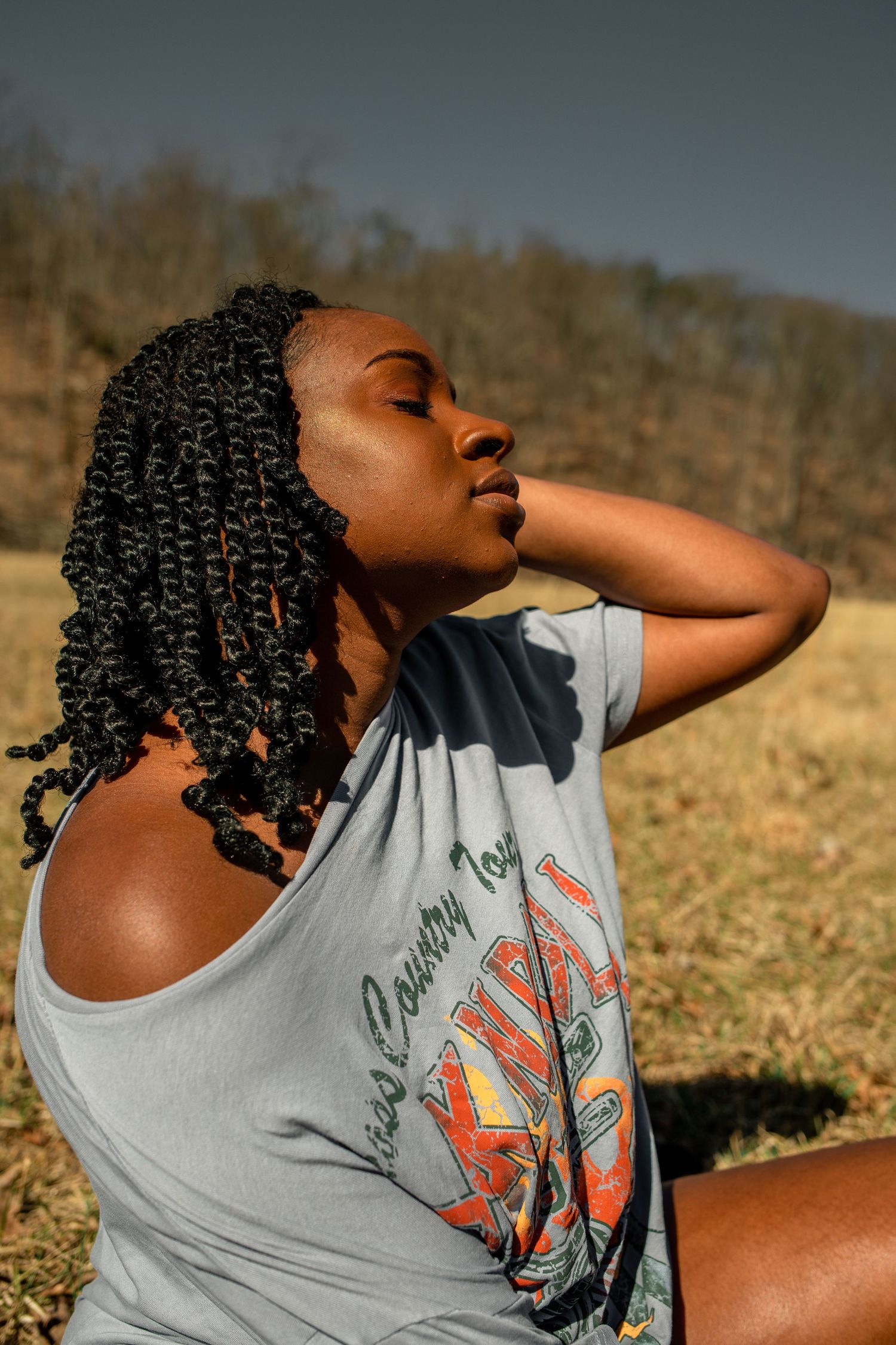 Young Black woman with braided hair poses outdoors in a graphic tee, eyes closed, hand behind neck.