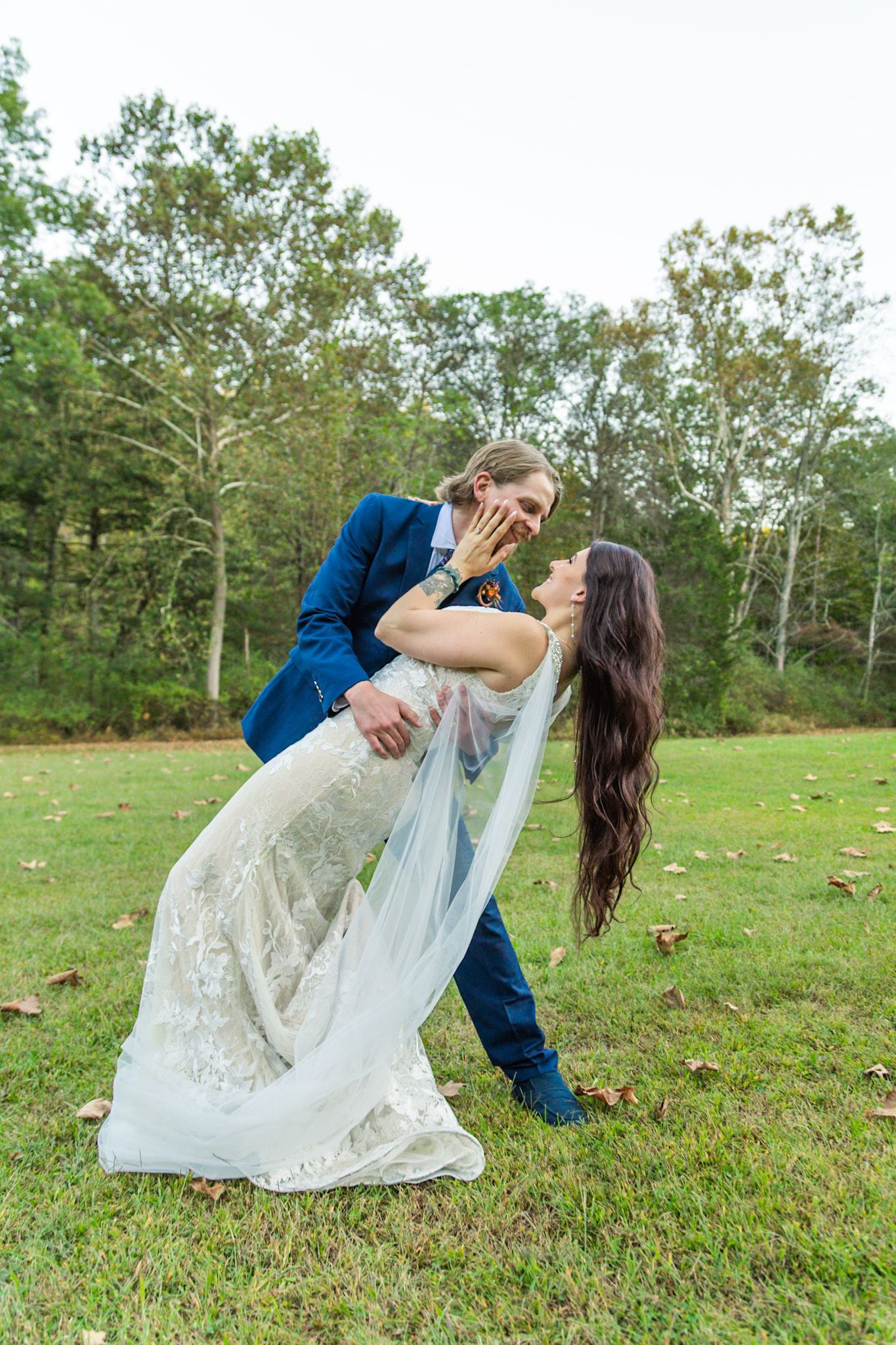 Groom dipping bride in lace wedding dress outdoors on green grass with trees in background.