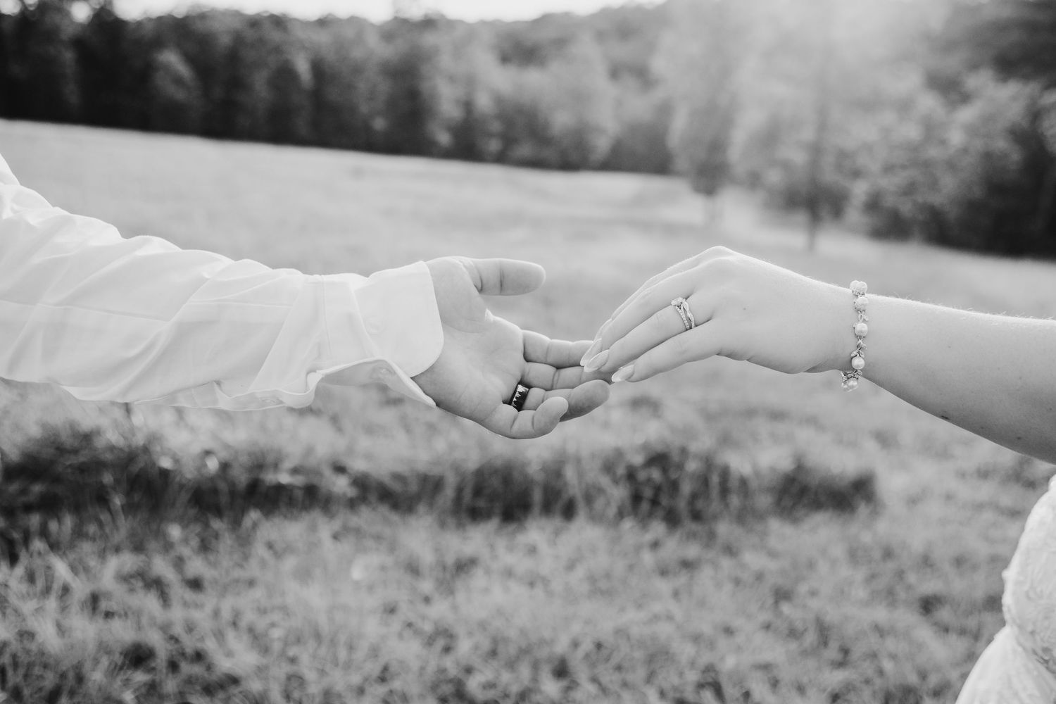 Black and white photo of a couple holding hands outdoors, showing wedding rings and a delicate bracelet.