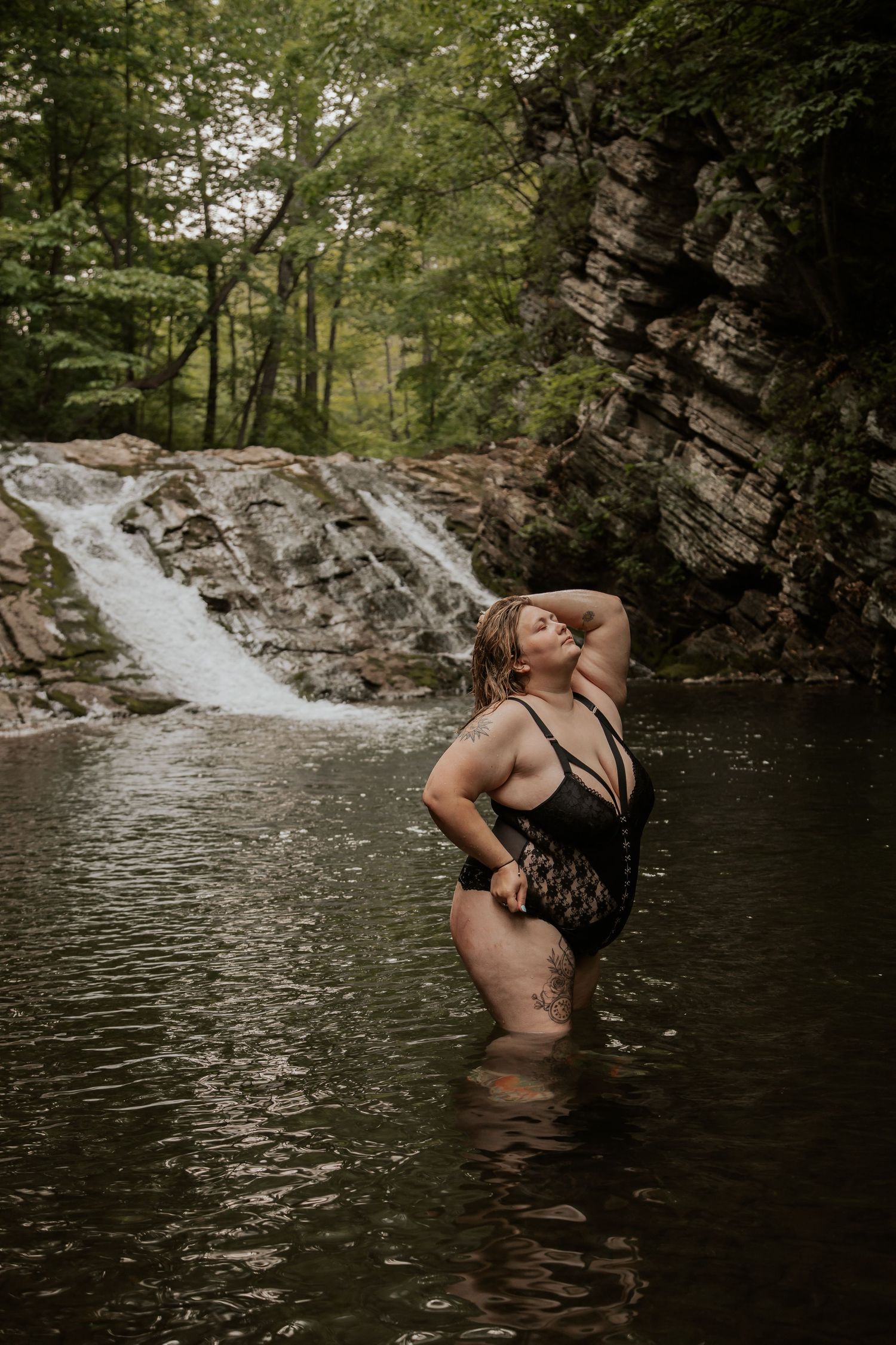 Plus-size woman in black lace swimsuit standing in a forest river with a waterfall behind her.