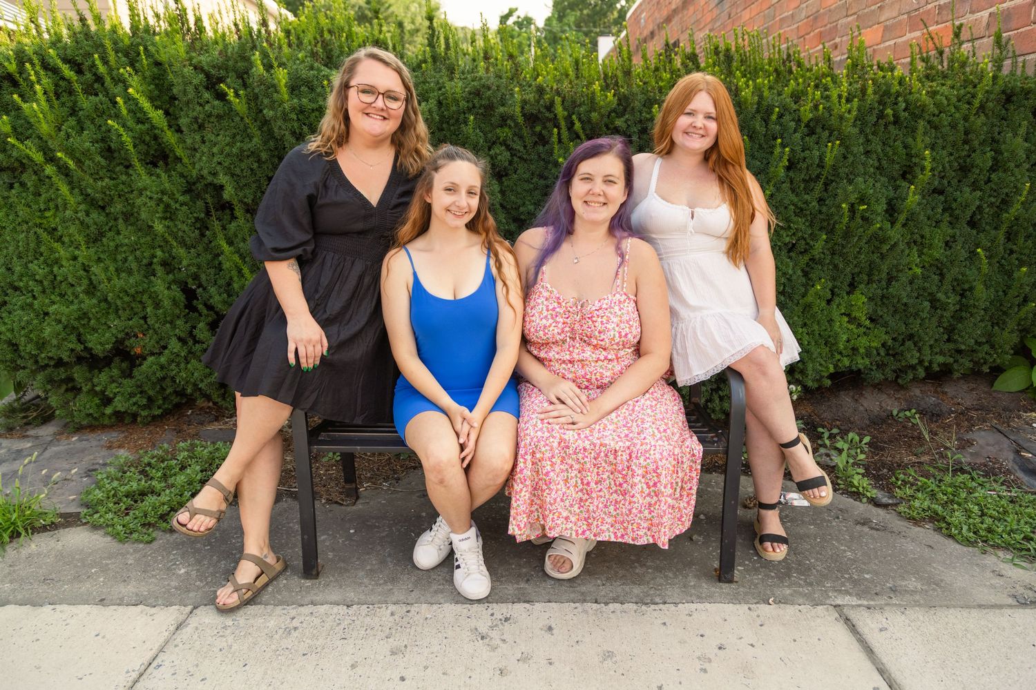 Four young women smiling together on a bench outdoors, surrounded by green hedges on a sunny day.