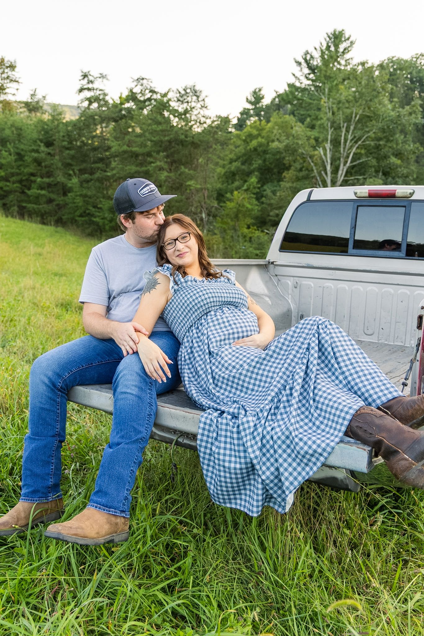 Expectant couple sitting on a pickup truck tailgate in a grassy field, woman in a blue gingham dress resting against the man in a cap.