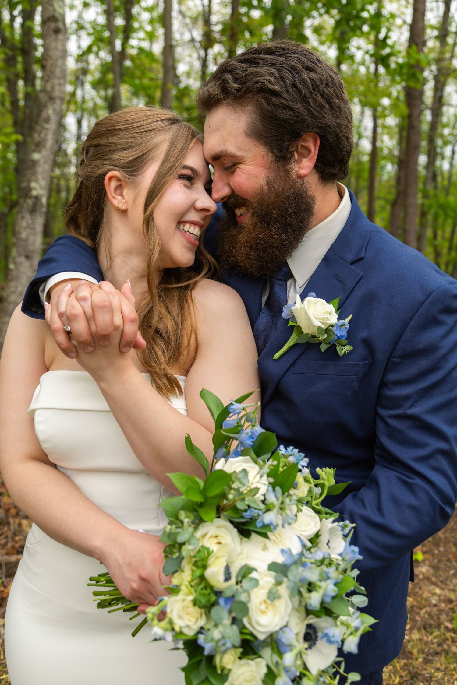 A smiling bride and bearded groom in a blue suit share a tender moment in a forest, holding a white and blue bouquet.