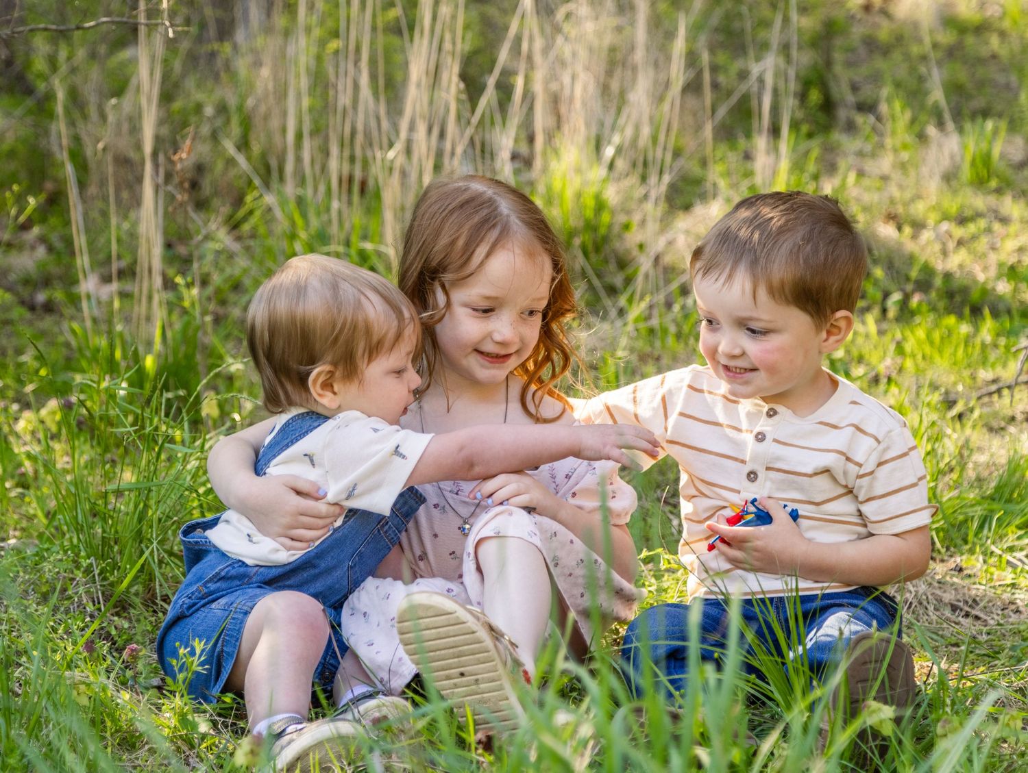 Three young children sitting together in green grass outdoors, with the older girl sharing berries with her siblings.