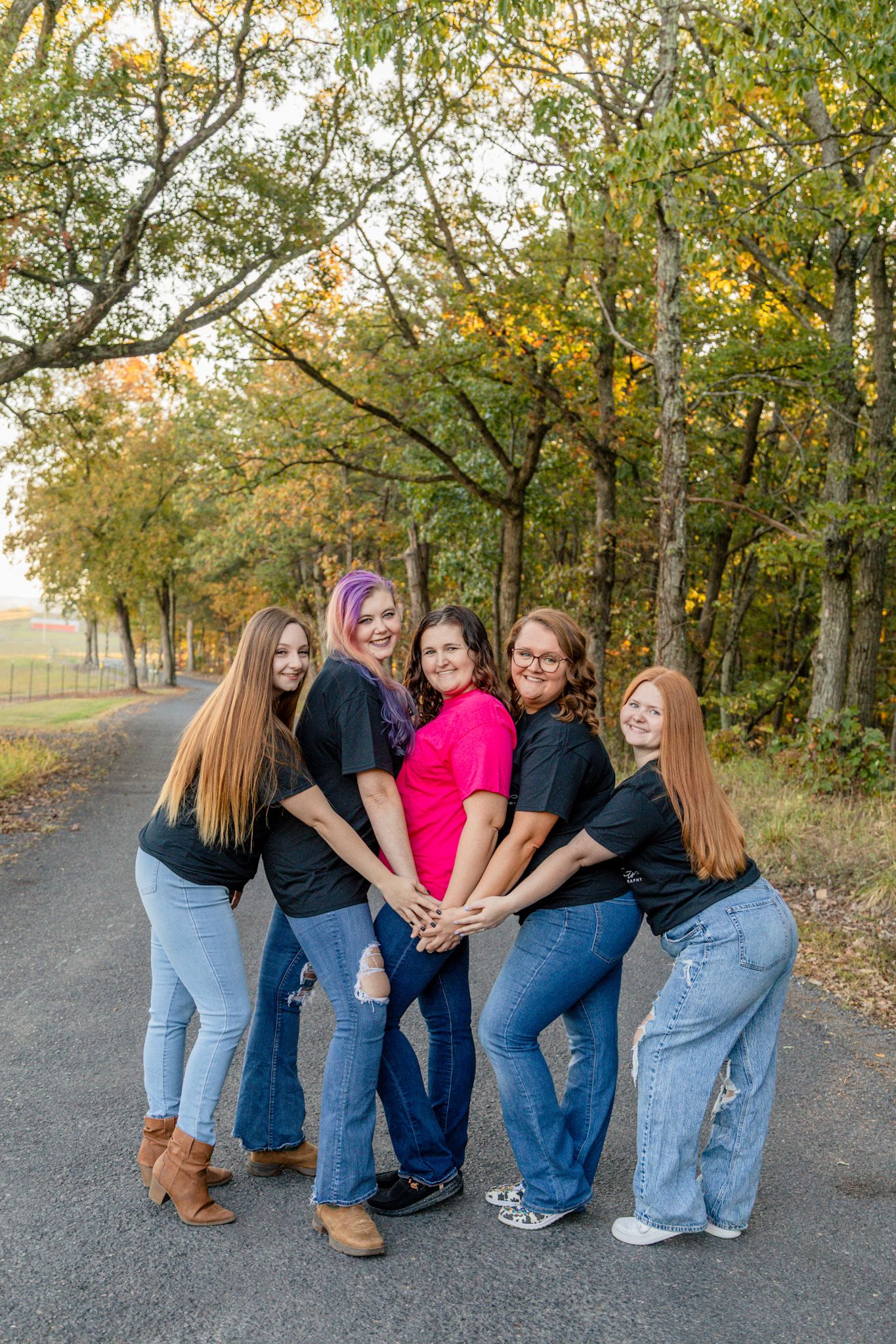 Five women in black and pink tops laughing together on a tree-lined road in autumn setting.