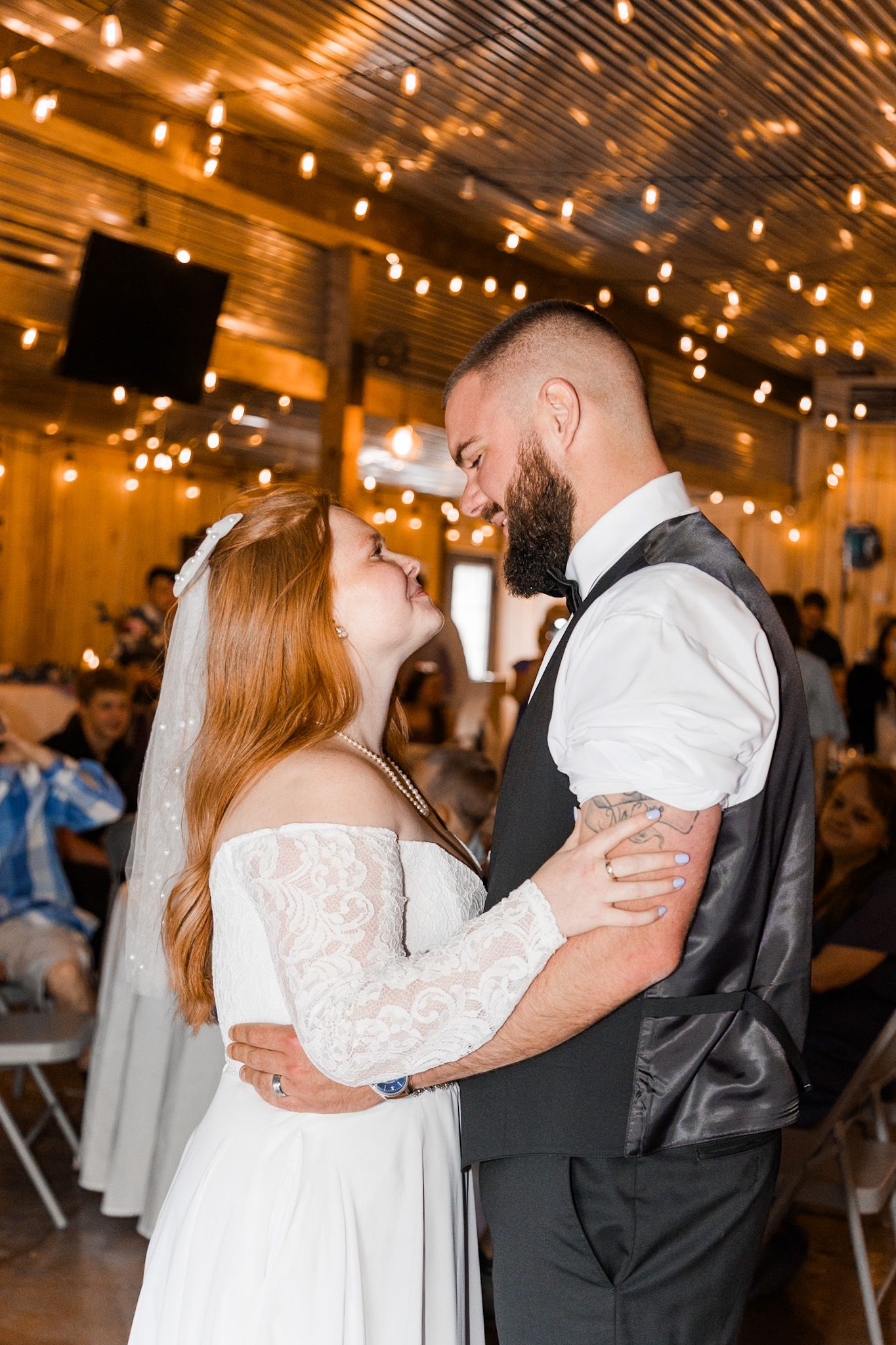 Bride and groom sharing first dance in rustic barn venue with warm string lights overhead.