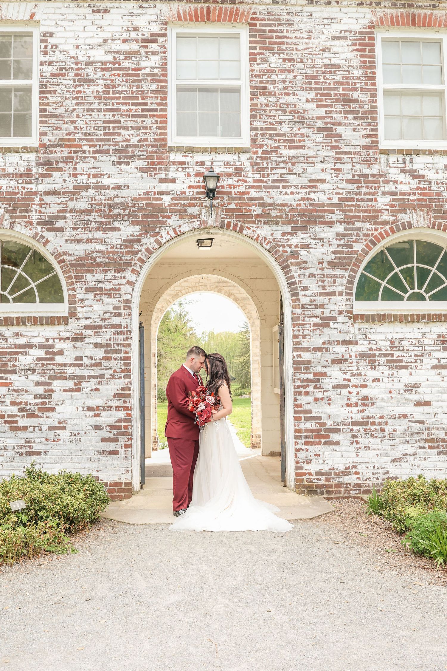 Bride and groom kissing under a brick archway holding a red floral bouquet at an elegant venue.