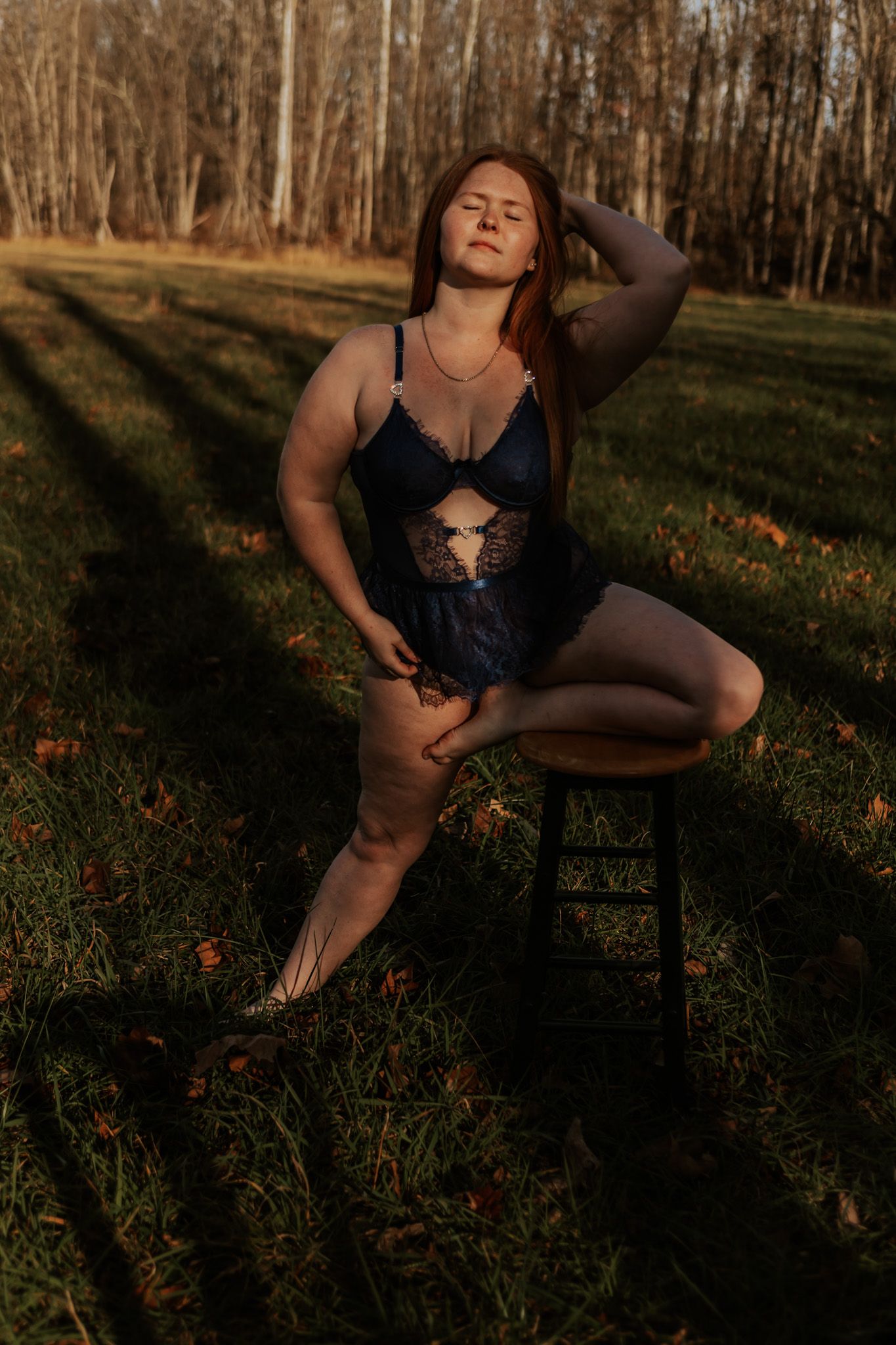 A redhead woman in navy blue lace lingerie poses on a stool in a sunlit outdoor setting.