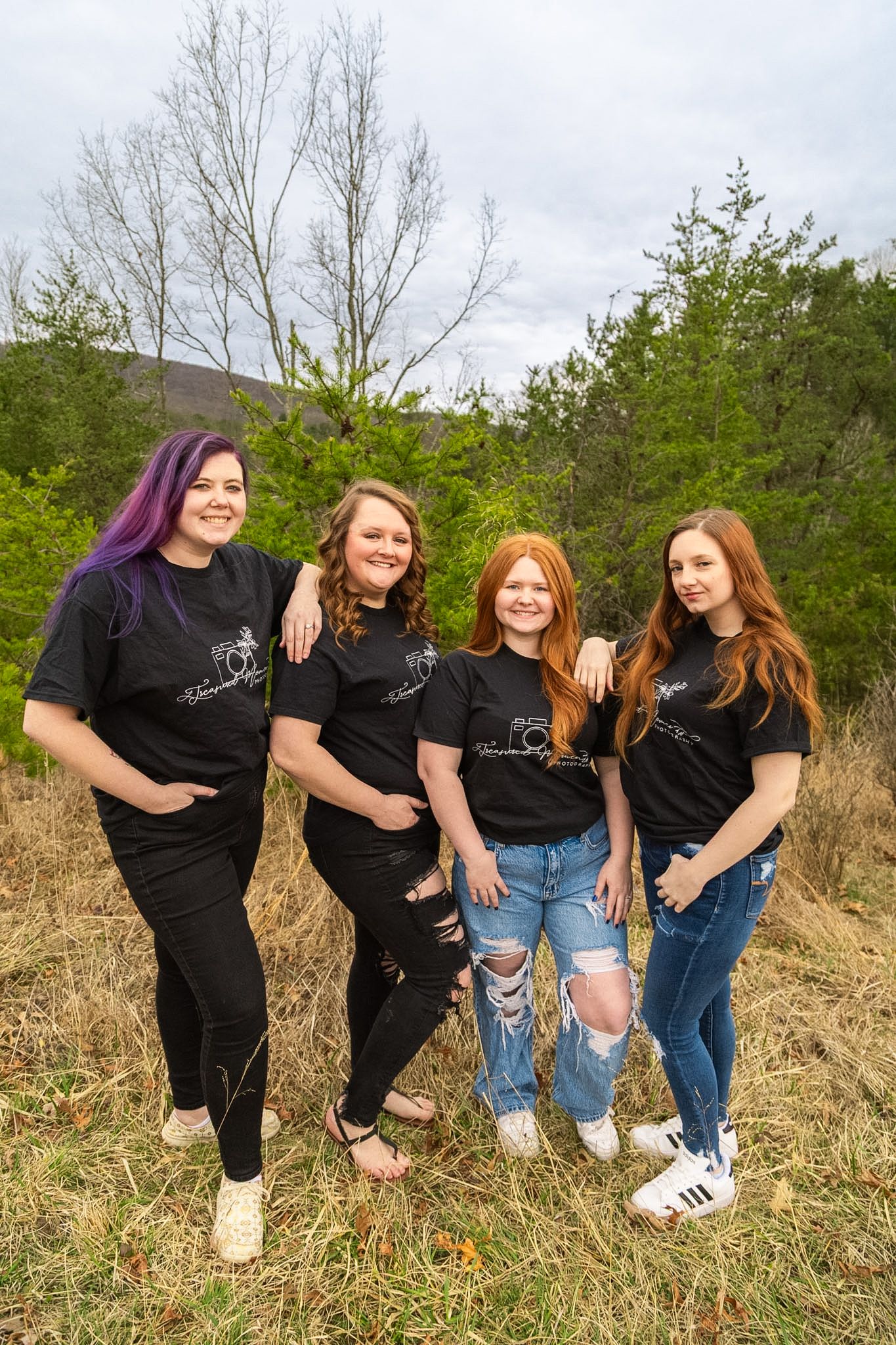 Four women in matching black Treasured Moments Photography shirts posing together in a field with trees behind them.