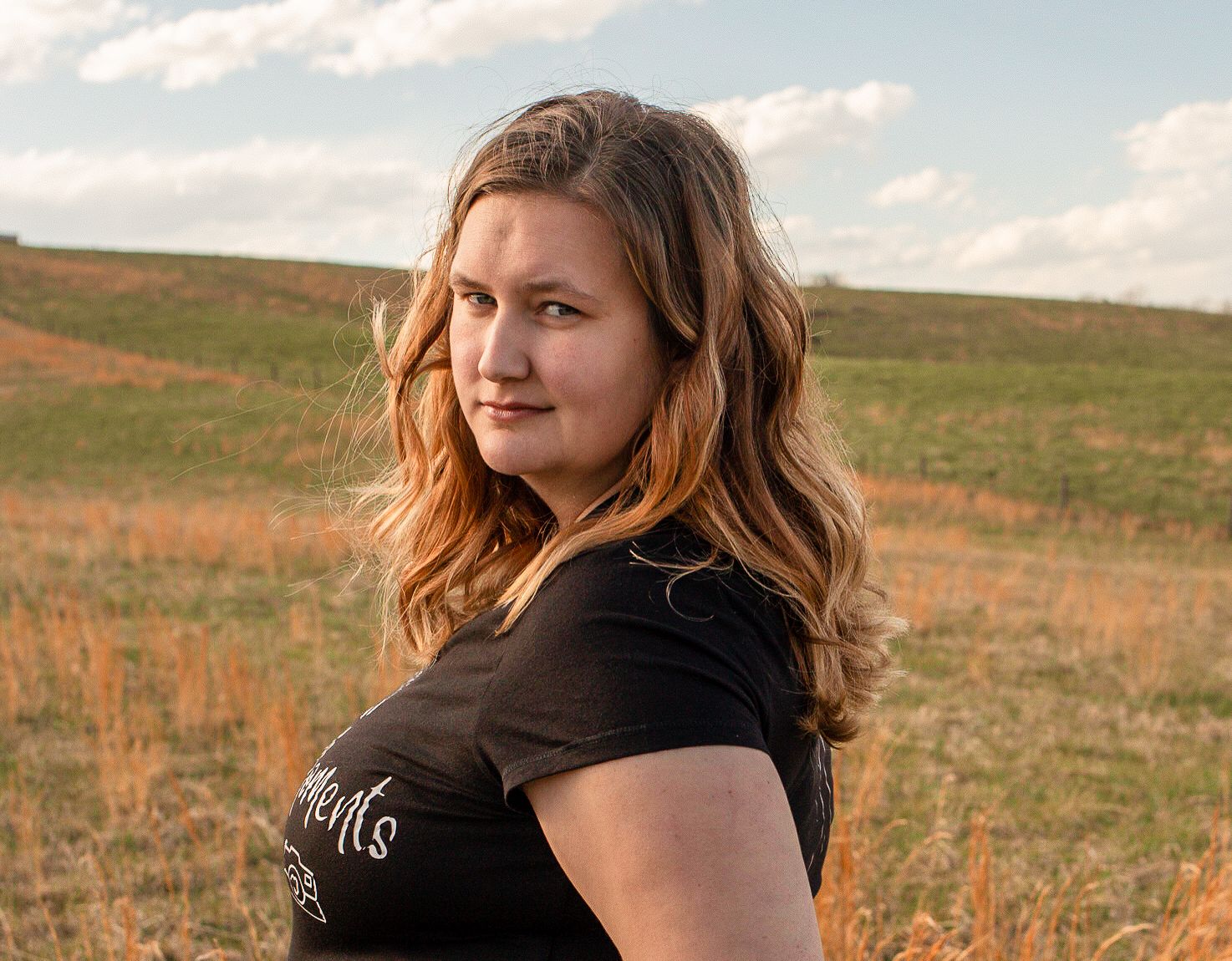Young woman with wavy hair in a black shirt glancing over her shoulder in a sunlit grassy field.