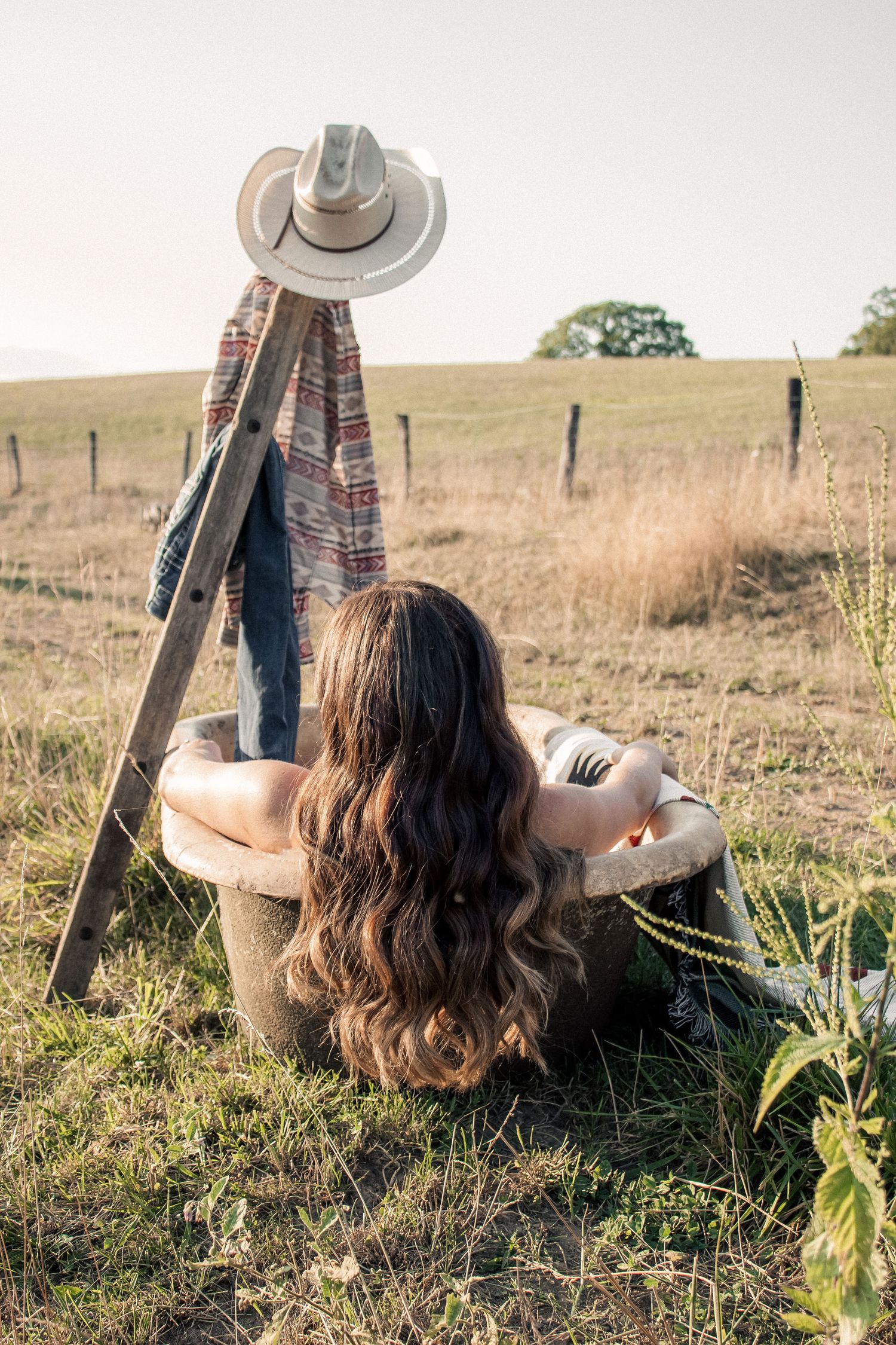 Woman with long wavy hair relaxing in a bathtub outdoors near a rustic ladder with cowboy hat in a field.