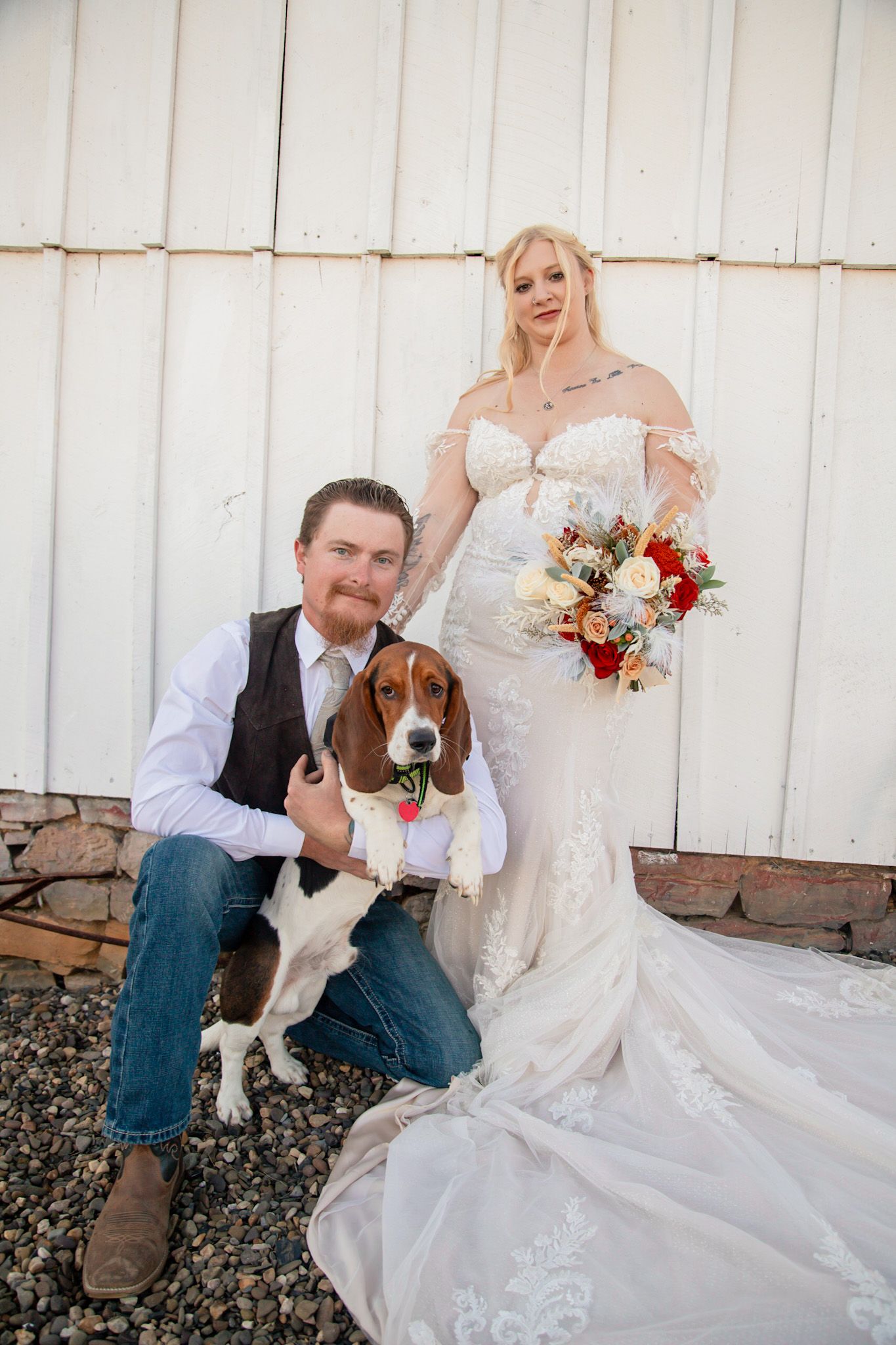 Wedding couple posing with beagle dog in bow tie against white barn wall with bouquet.