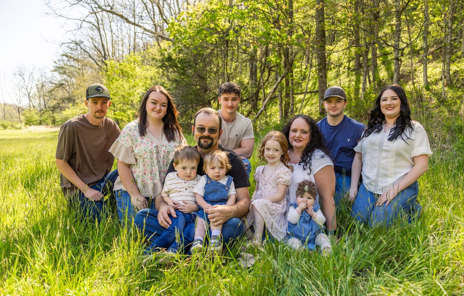 Large family group portrait outdoors in lush green meadow with trees, smiling together on a sunny spring day.