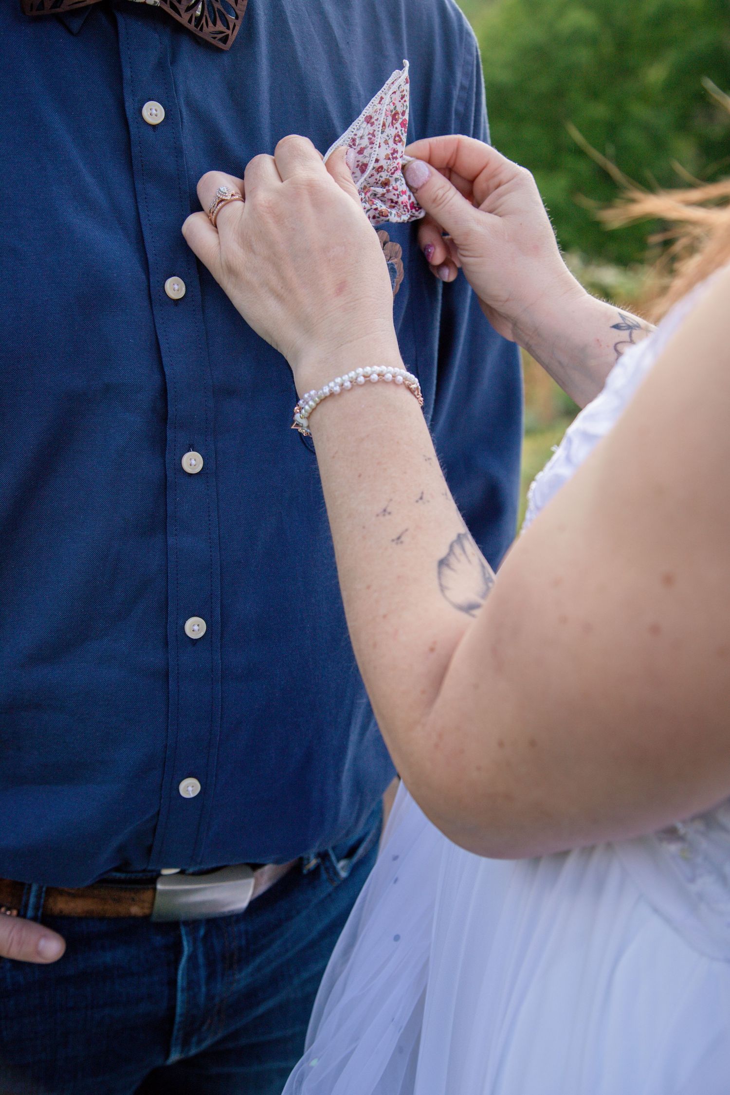 Bride with pearl bracelet and tattoo adjusting floral pocket square on groom in blue shirt.
