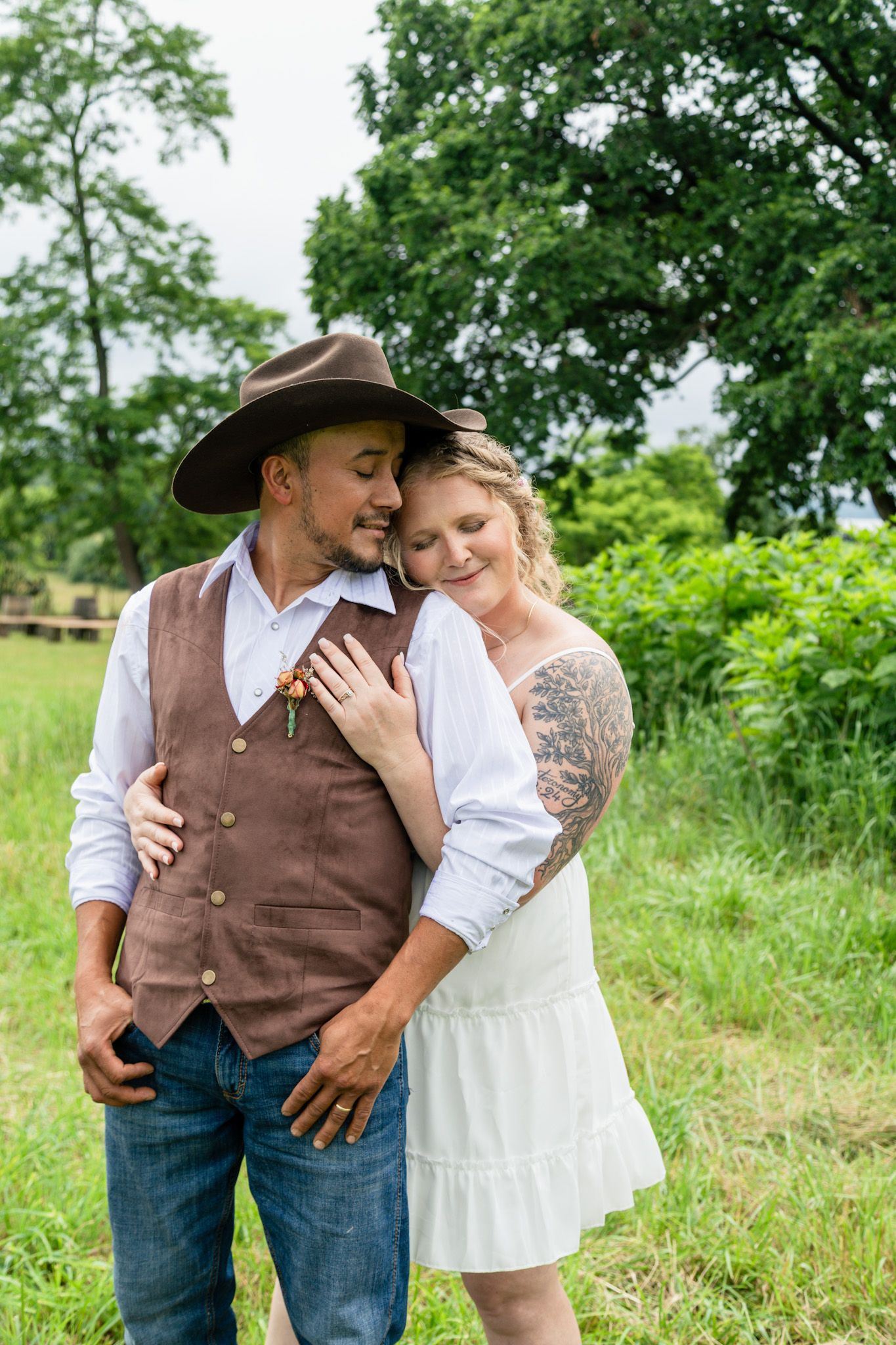 Couple embracing outdoors, man in brown vest and cowboy hat, woman in white dress with tattoos.