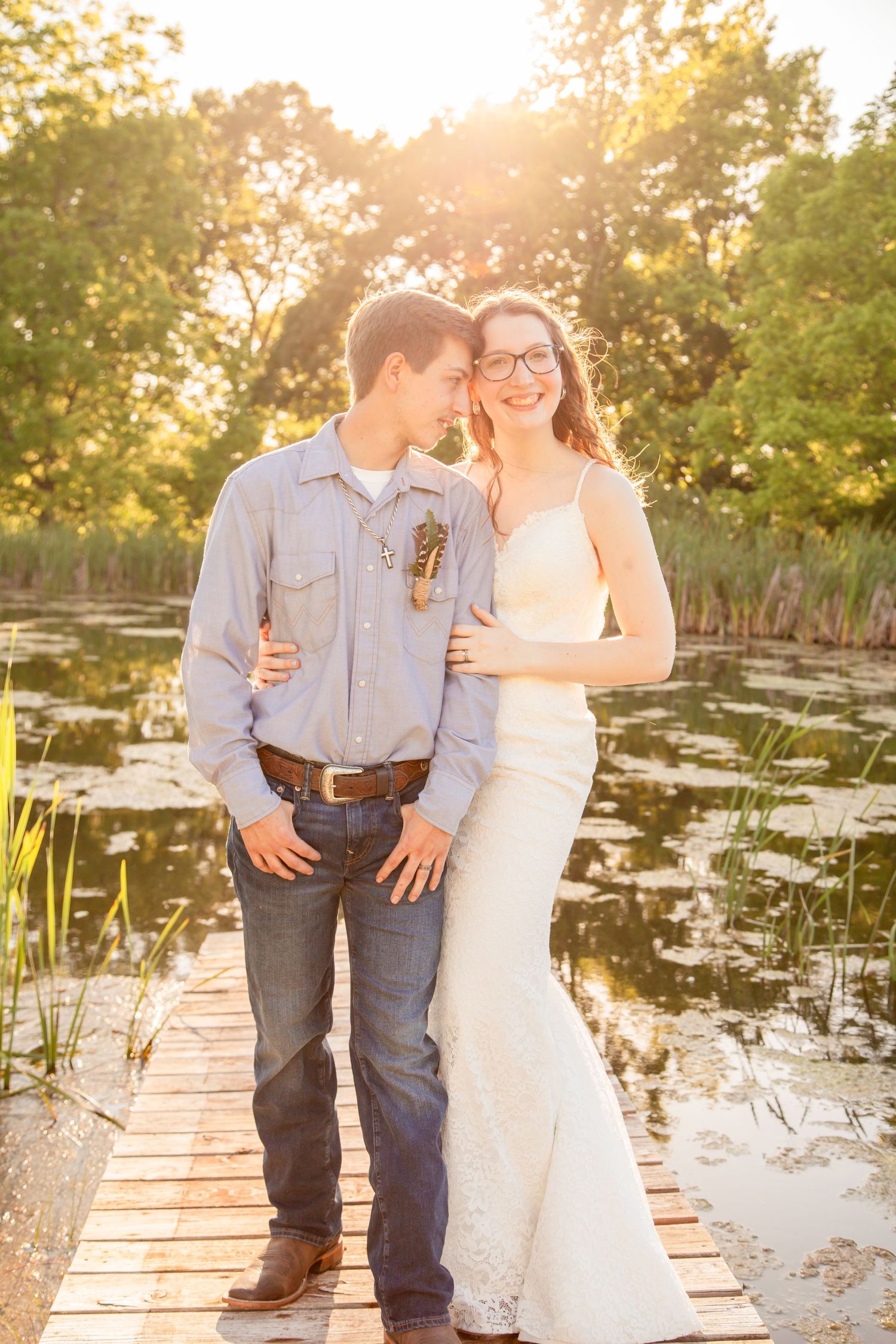 Couple sharing a romantic moment on wooden dock at sunset by a serene pond surrounded by trees.