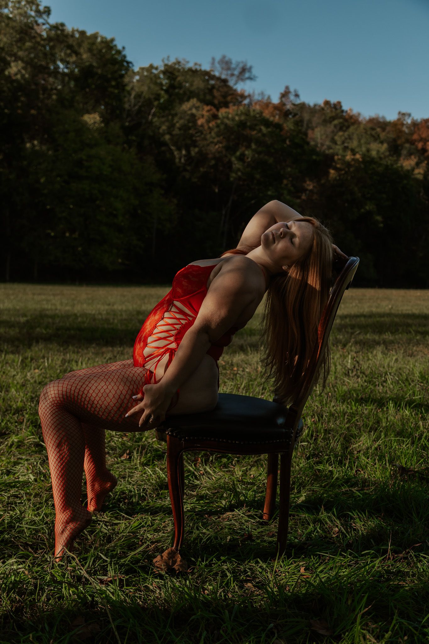 A woman in red lingerie and fishnet stockings poses on a vintage chair in an autumn field.