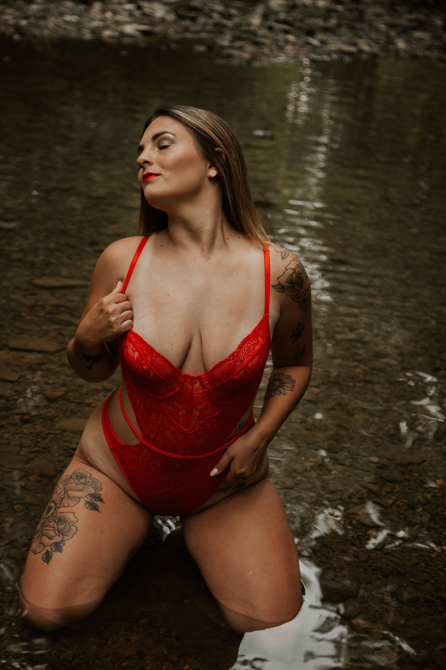 Woman in red lace bodysuit kneeling in a shallow creek with tattoos visible on arms and thigh.