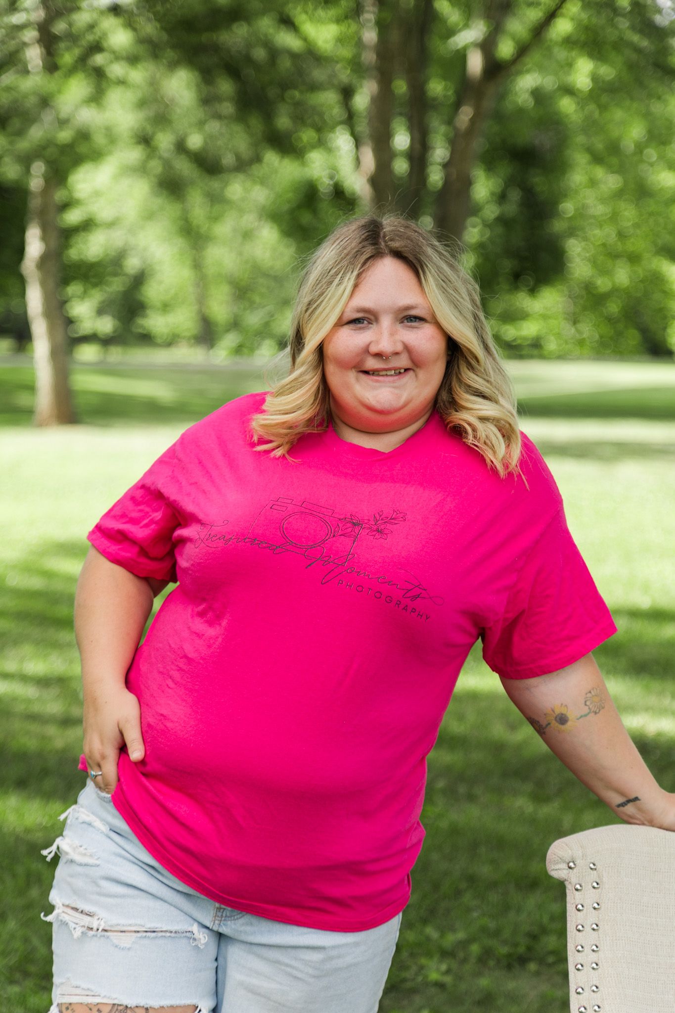 Smiling blonde woman in bright pink t-shirt and ripped jeans posing outdoors in a lush green park.