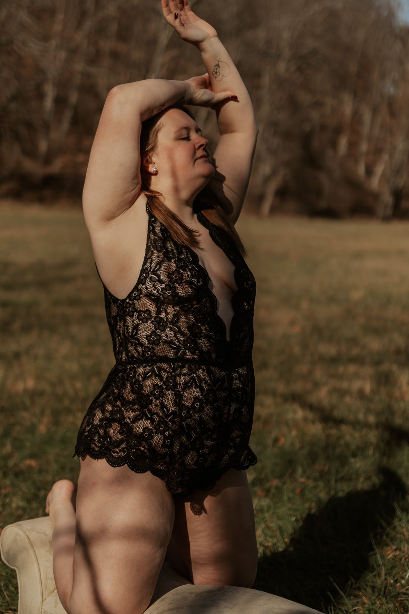 Woman in black lace bodysuit kneels with arms raised in sunlit outdoor autumn meadow.
