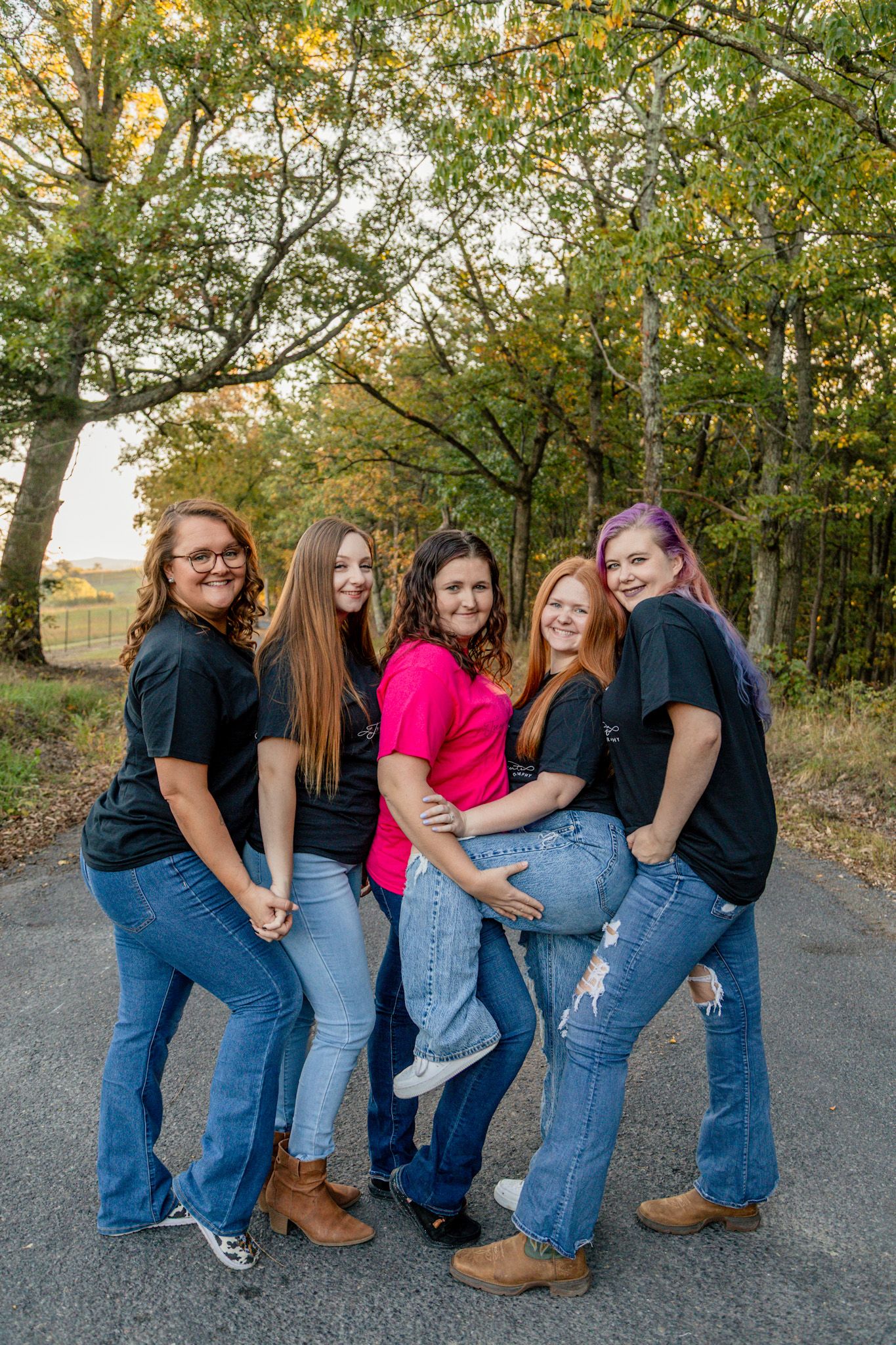 Group of five women posing together on a tree-lined road wearing black and pink shirts and jeans.