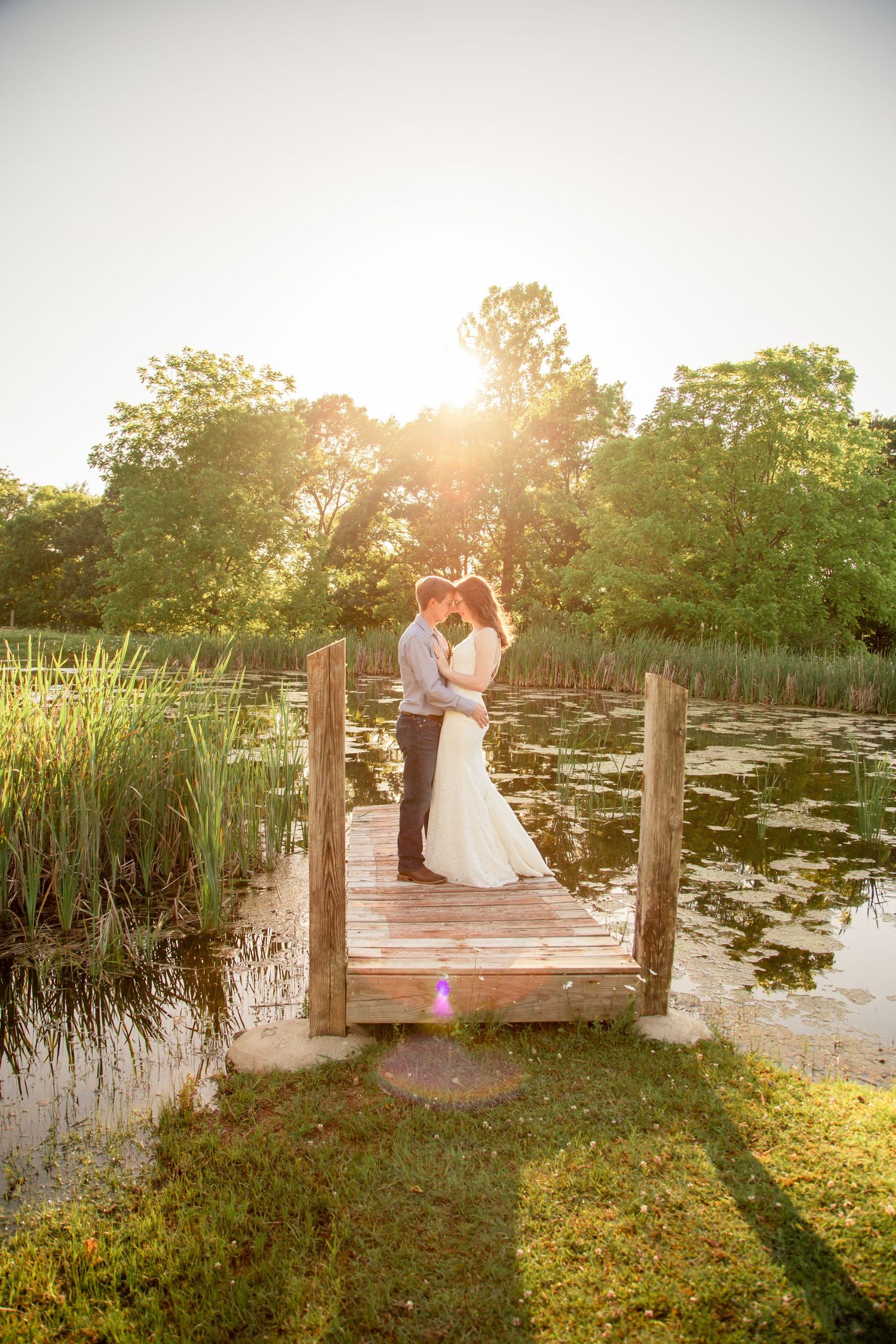Newlyweds embrace on a wooden dock at sunset surrounded by a serene pond and lush greenery.
