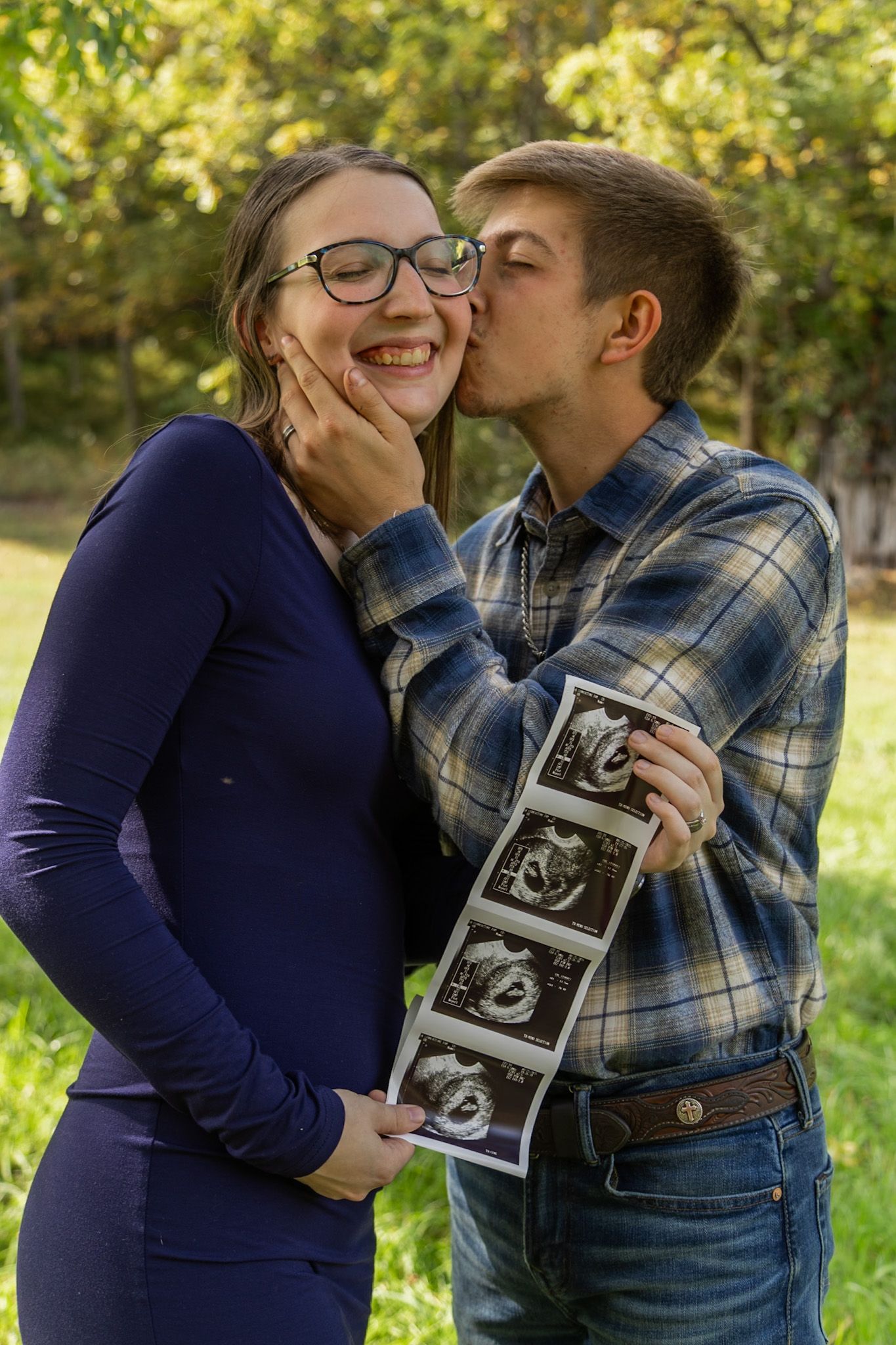 Happy pregnant couple sharing a kiss outdoors, holding ultrasound photos while surrounded by green trees.
