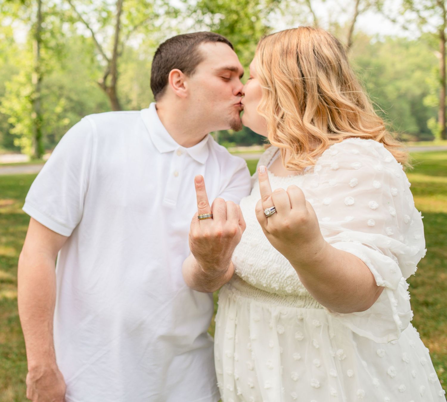 Couple kissing while showing off wedding rings in sunny outdoor park setting.