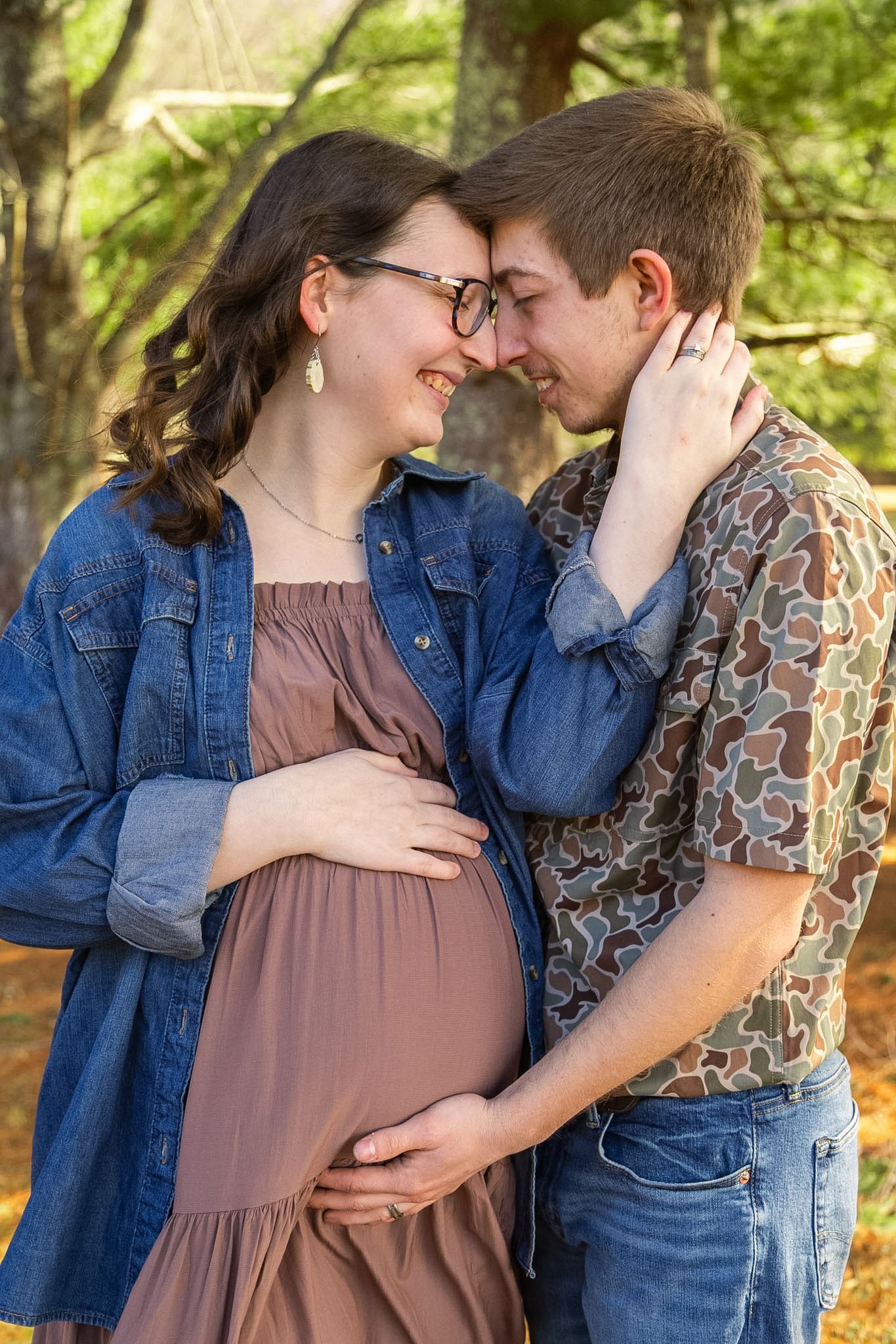 Smiling pregnant couple touching foreheads tenderly in a sunlit autumn forest, woman in denim jacket and mauve dress.