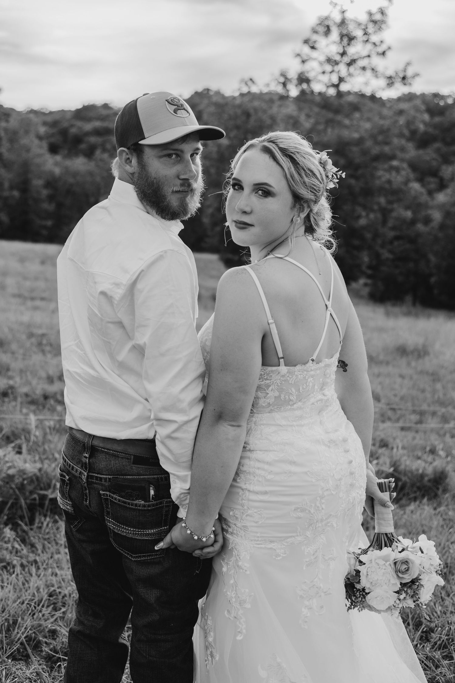 Black and white photo of a bride and groom holding hands in a field, bride in lace gown holding bouquet.