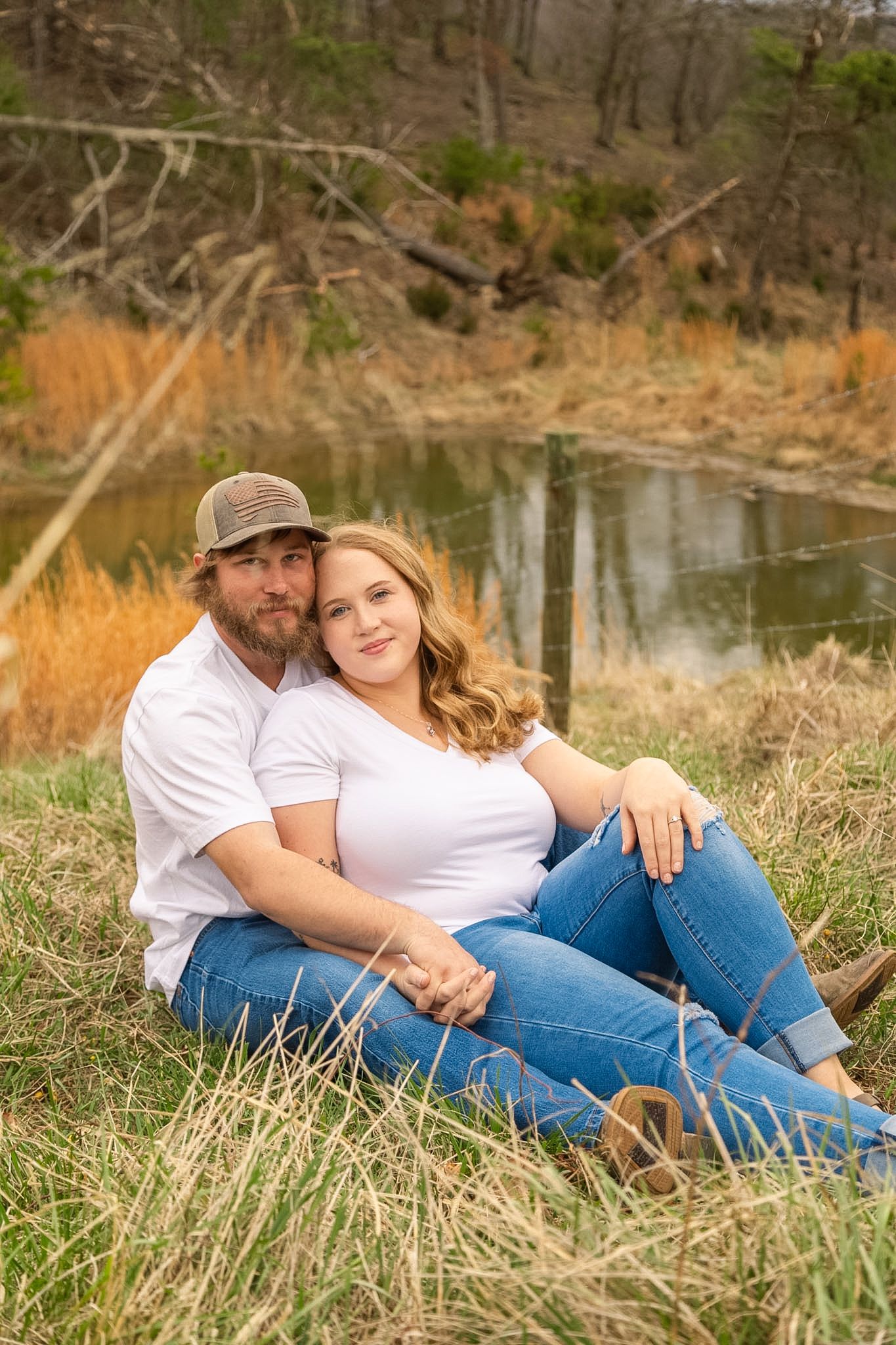 Couple sitting together on grass near a pond, smiling warmly, both wearing white shirts and jeans.