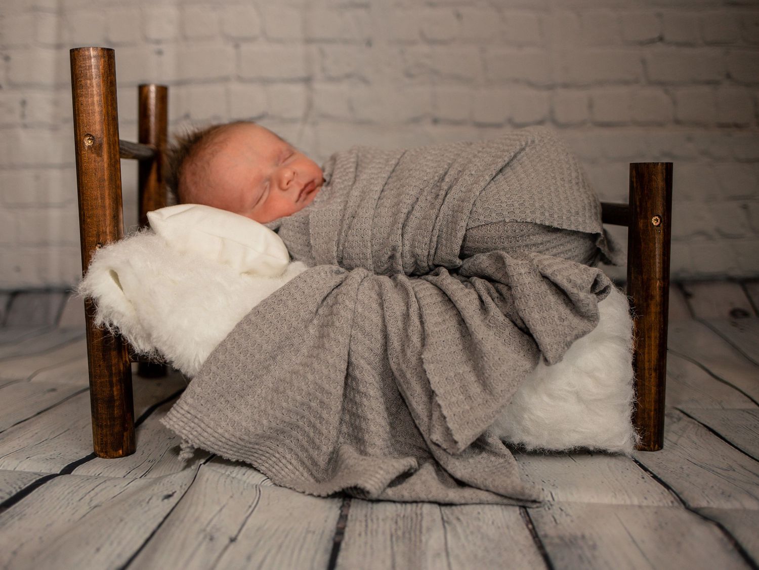 Sleeping newborn baby wrapped in a gray knit blanket on a rustic wooden miniature bed.