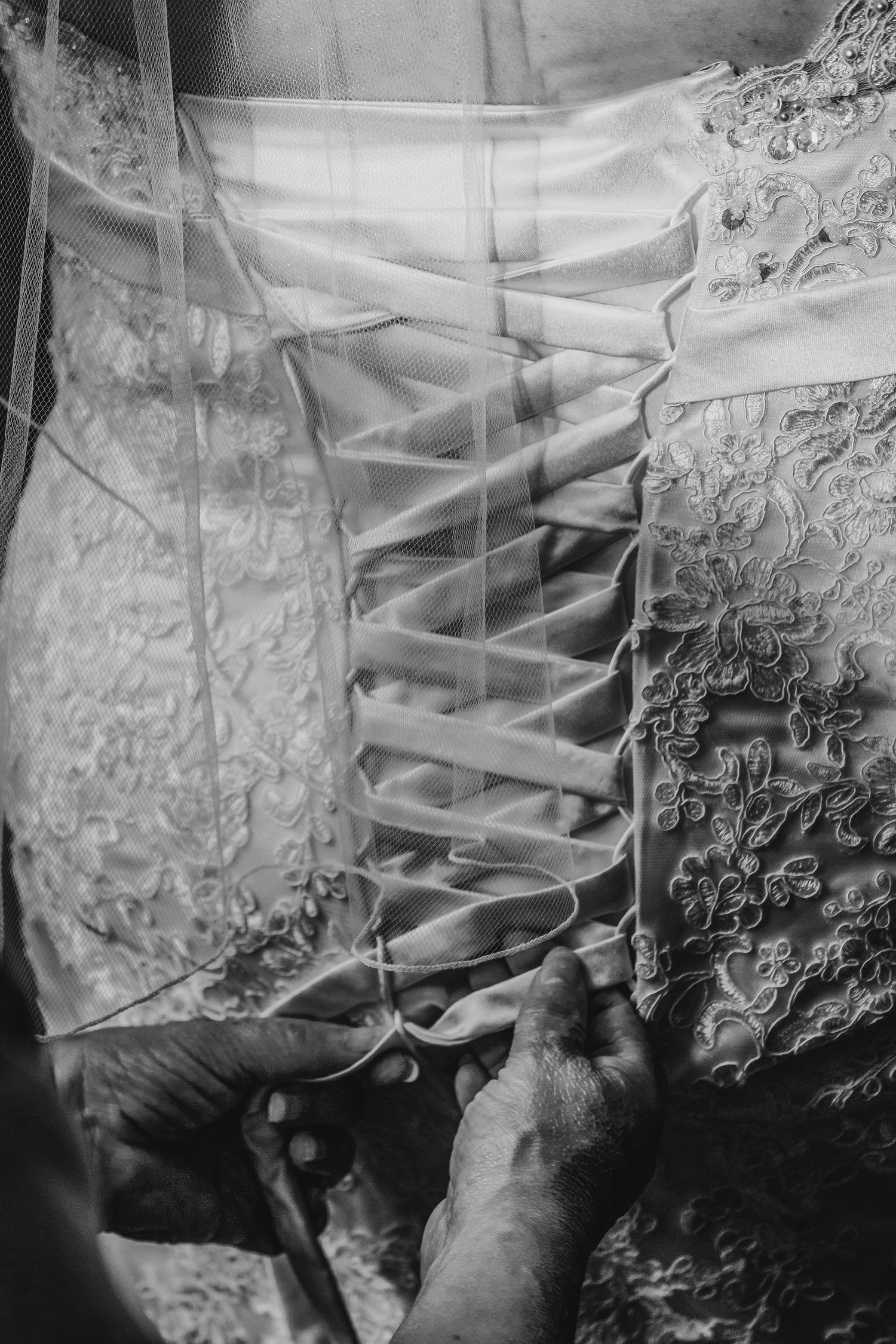 Black and white close-up of hands lacing up corset back of elegant lace wedding gown with veil.